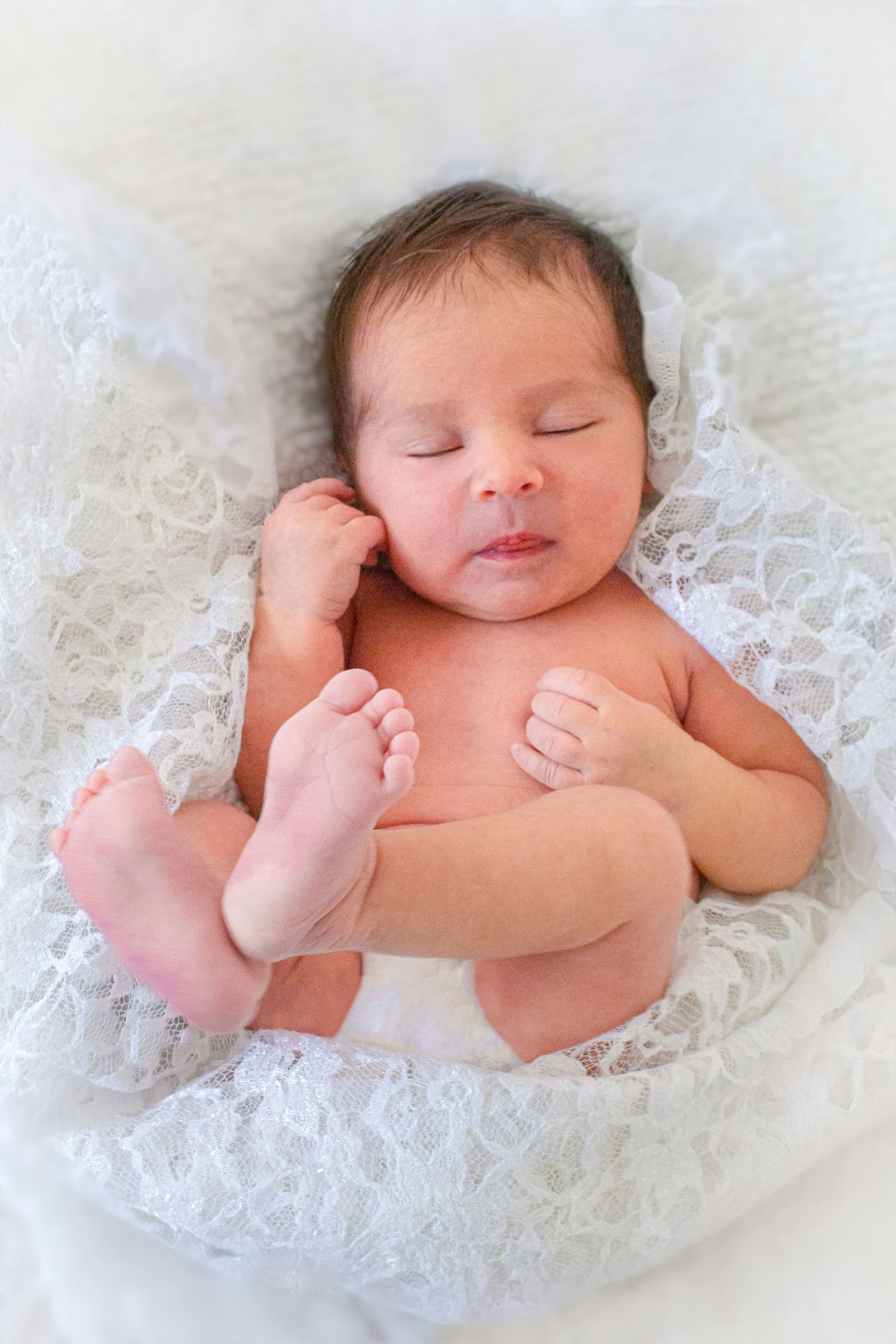 Newborn baby sleeping peacefully in a white lace nest with tiny feet visible.