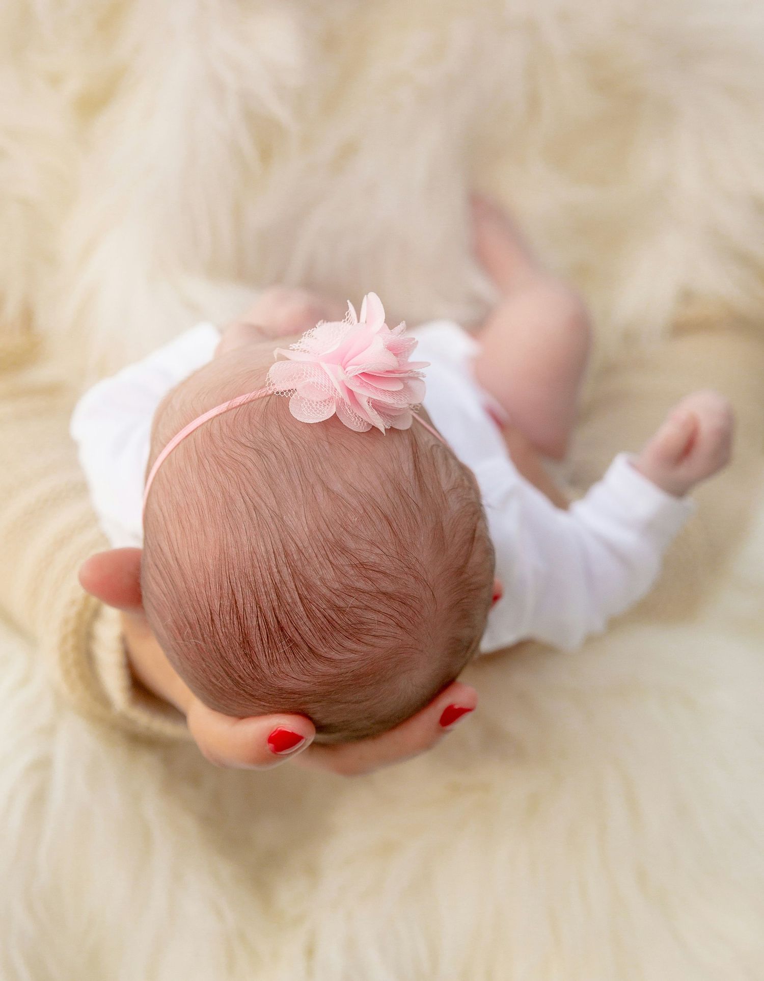 Newborn baby with pink floral headband lying on cream fur blanket.