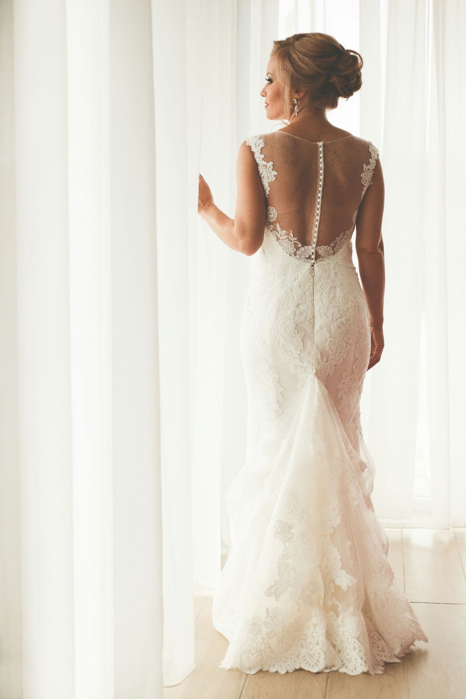 Bride in fitted lace wedding dress with illusion back and button detail standing by curtains.