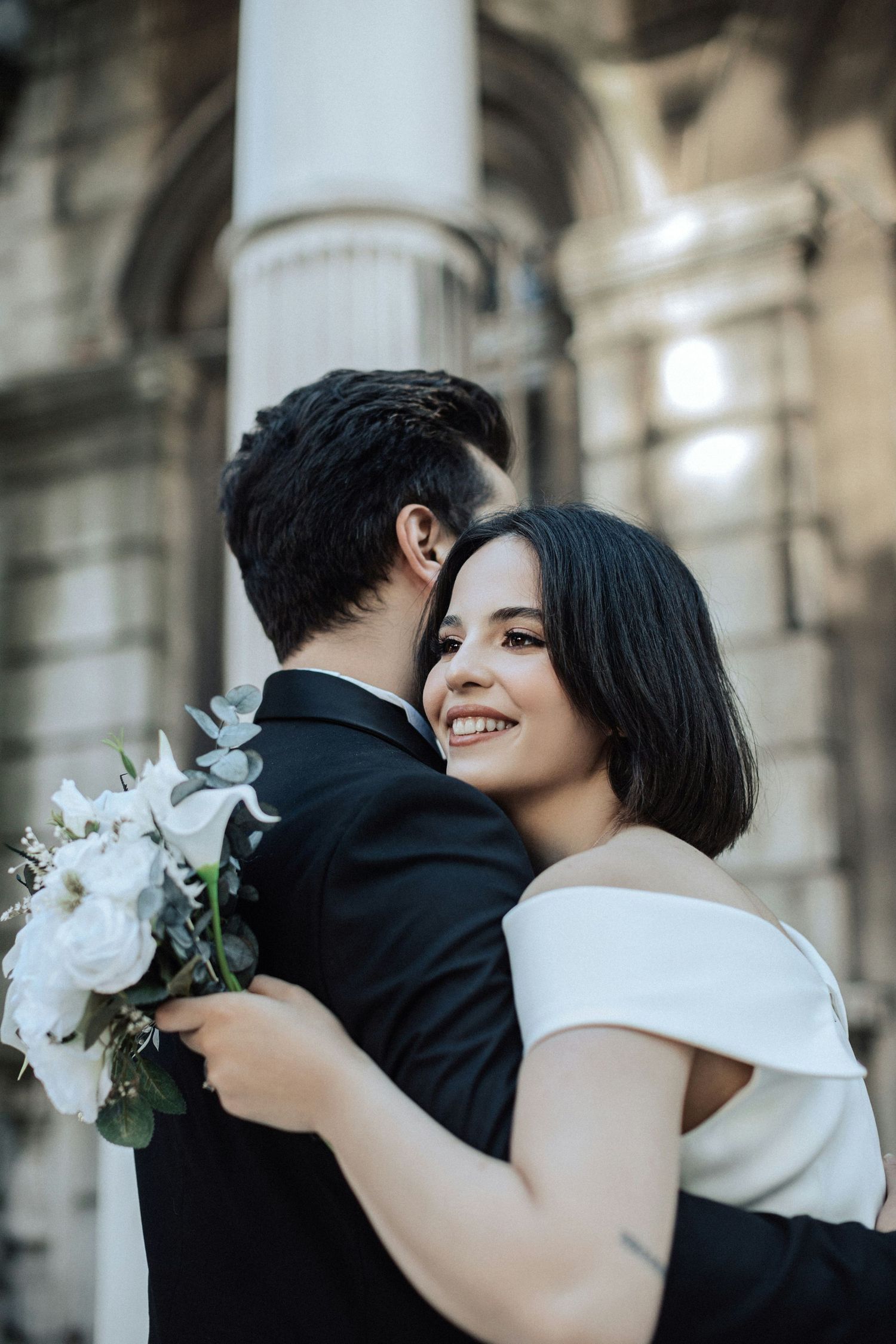 Smiling dark-haired bride in off-shoulder white gown embraces groom while holding white floral bouquet outdoors.