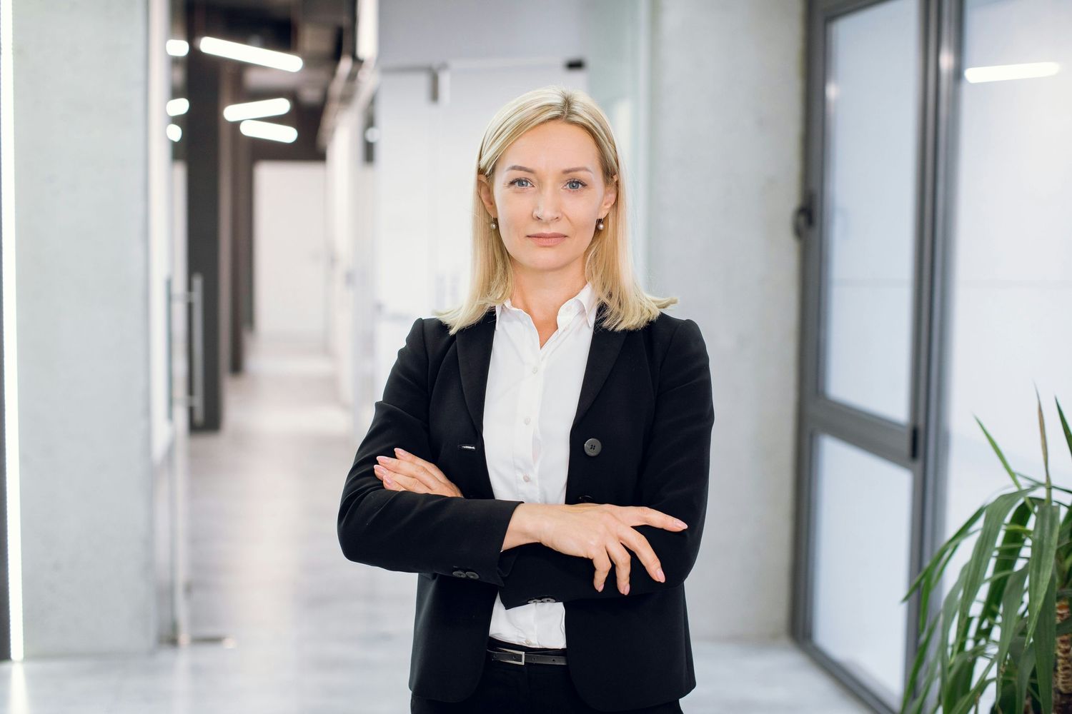 Professional businesswoman in black blazer stands confidently with arms crossed in a modern office hallway.