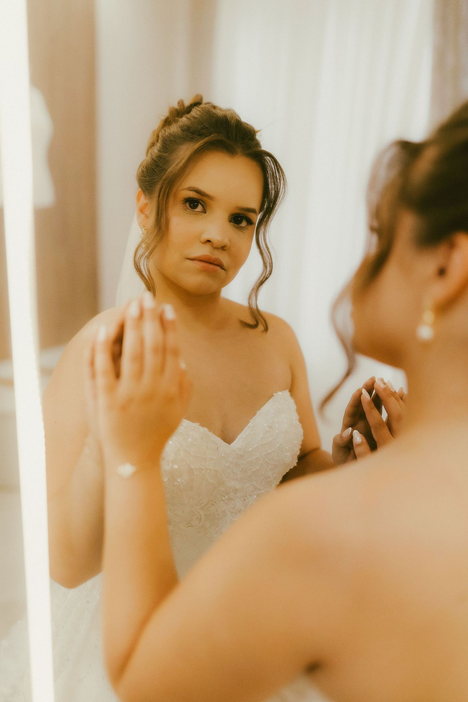 Bride in white strapless lace gown gazes at her reflection in a mirror, wearing elegant pearl earrings and bracelet.