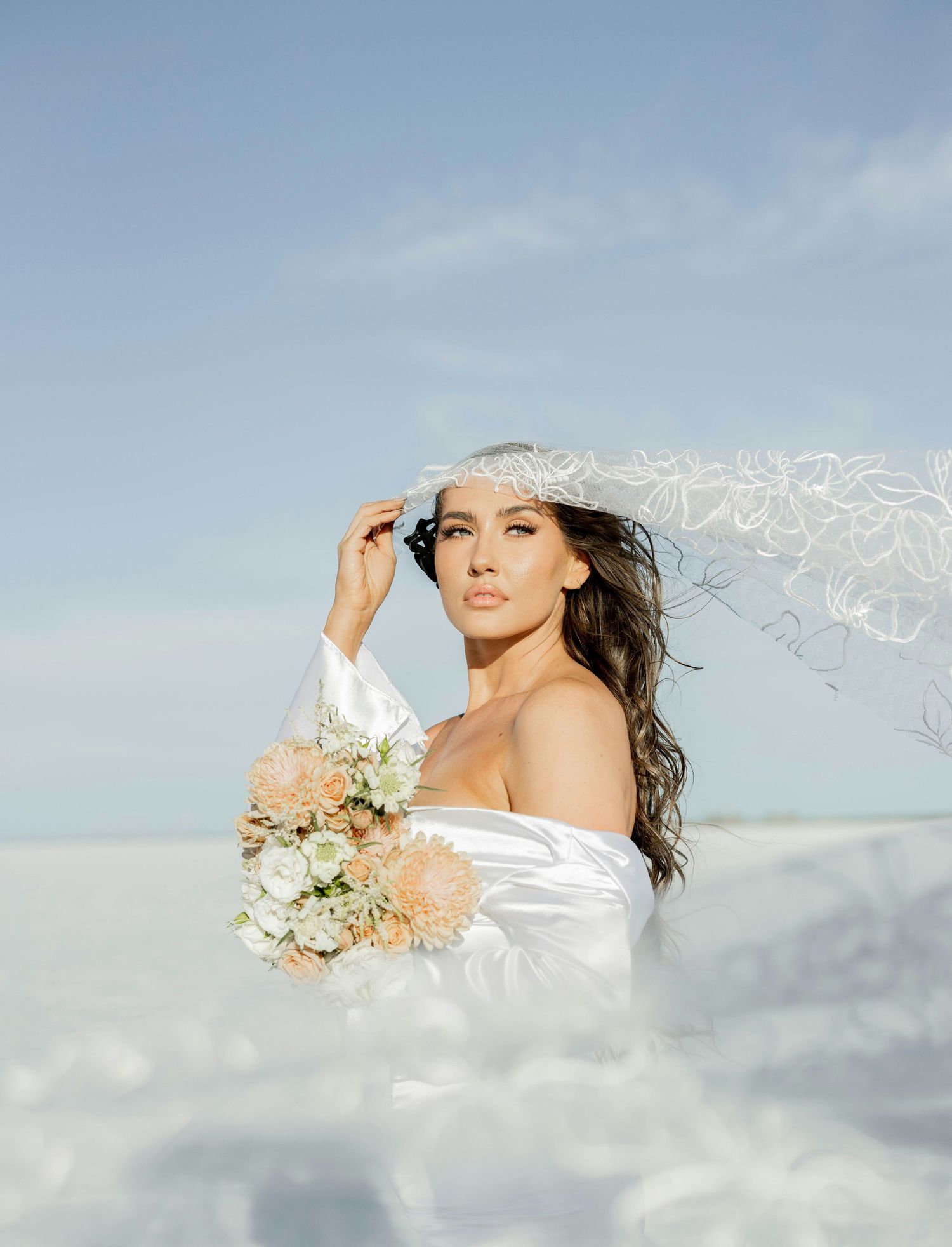 Elegant bride in white off-shoulder gown holding peach bouquet with flowing veil on sunny beach.