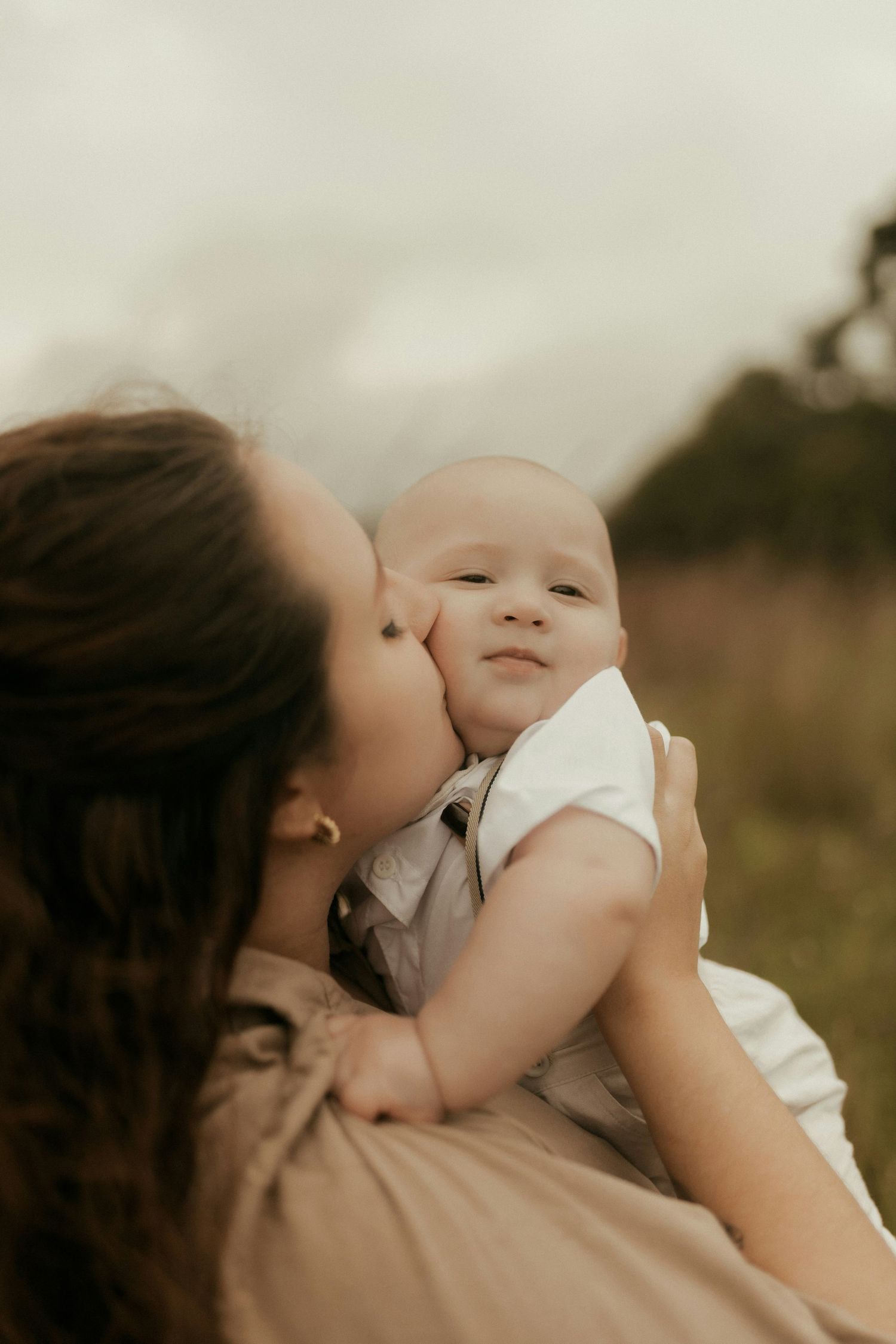 Mother kissing chubby-cheeked baby outdoors in green field on cloudy day.