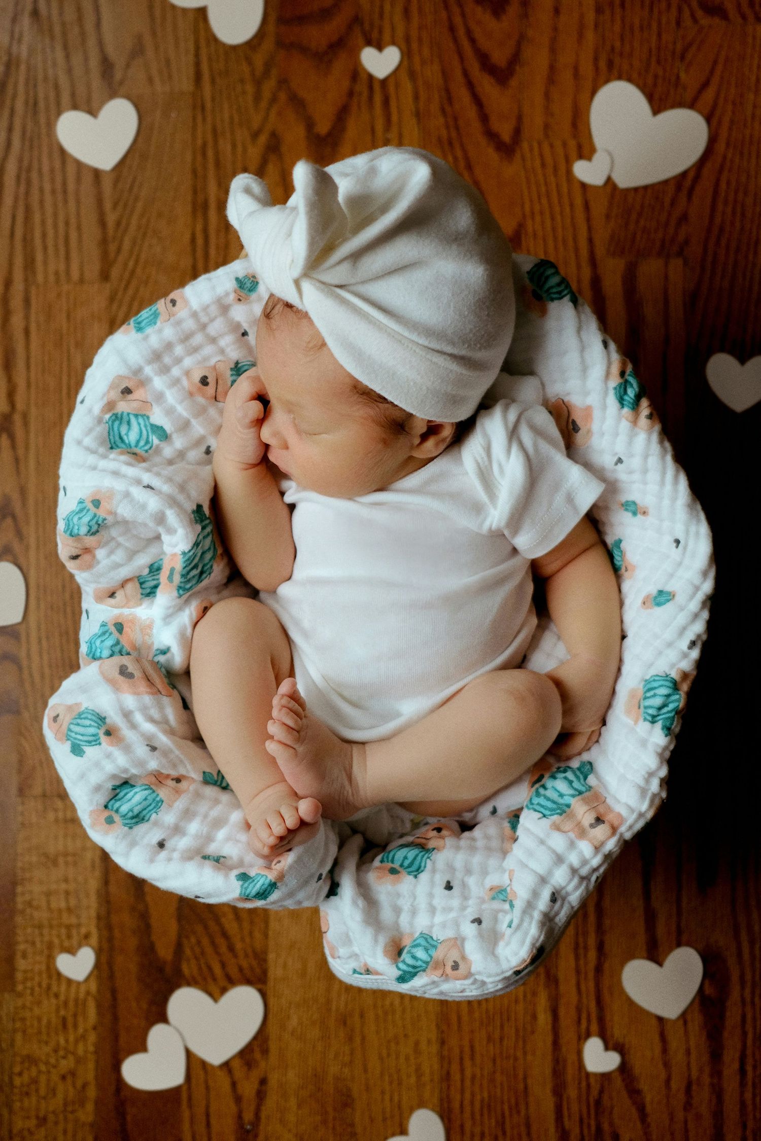 Newborn baby in white onesie and bow headband sleeping in a floral muslin nest on a wooden floor.