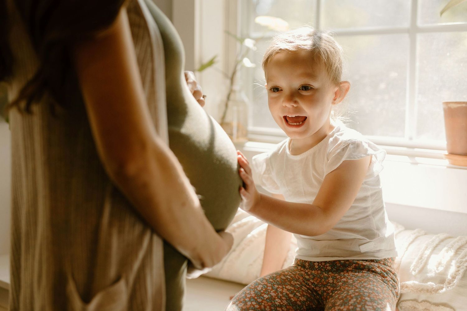 Excited toddler touching pregnant mothers belly while sitting by a sunny window at home.