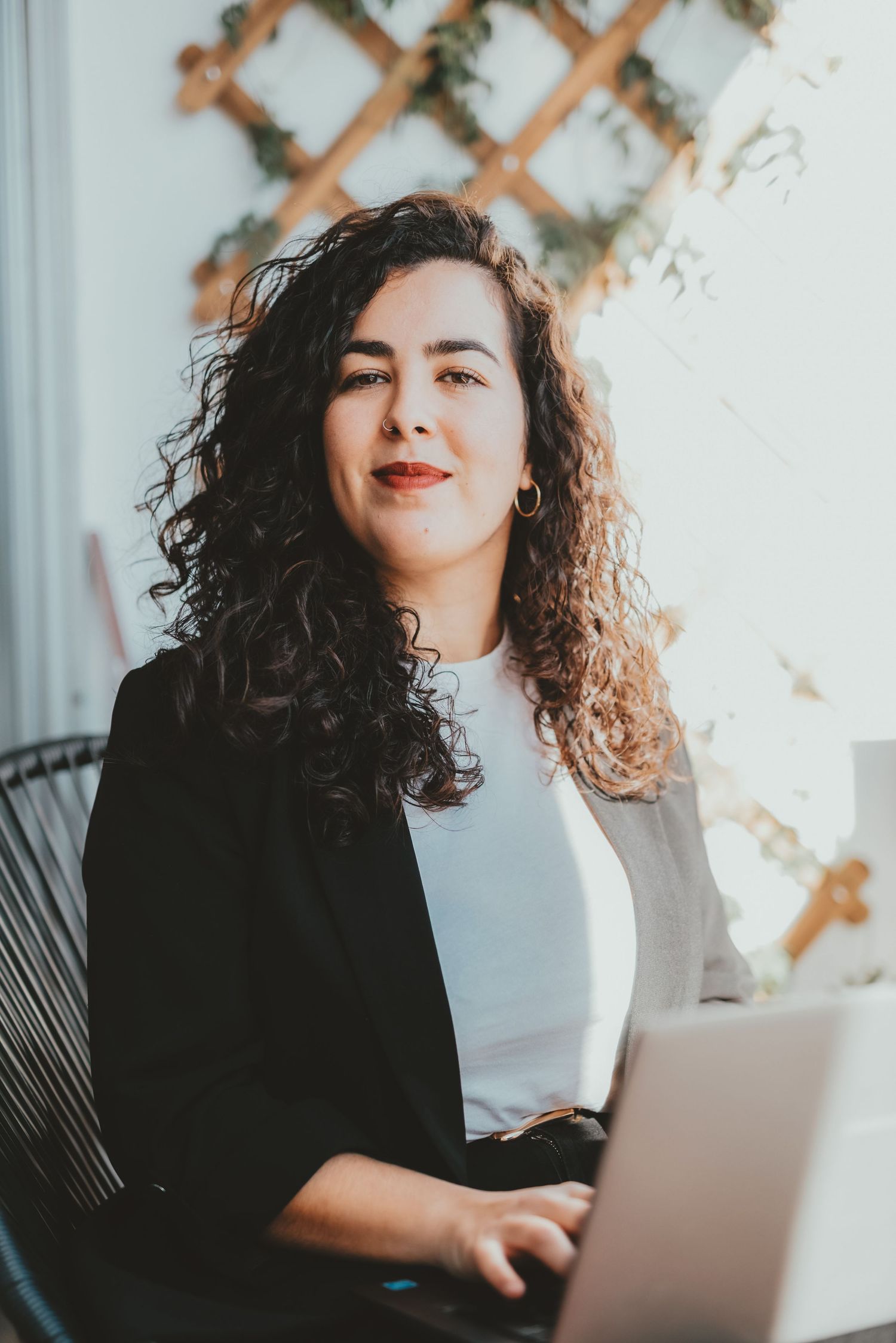 Confident curly-haired businesswoman smiling while using laptop in bright modern workspace.