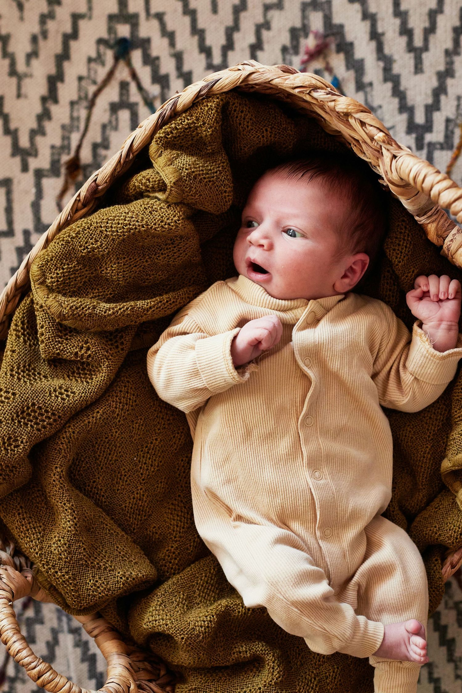 Alert newborn baby lying in a wicker basket wrapped in an olive green knit blanket.