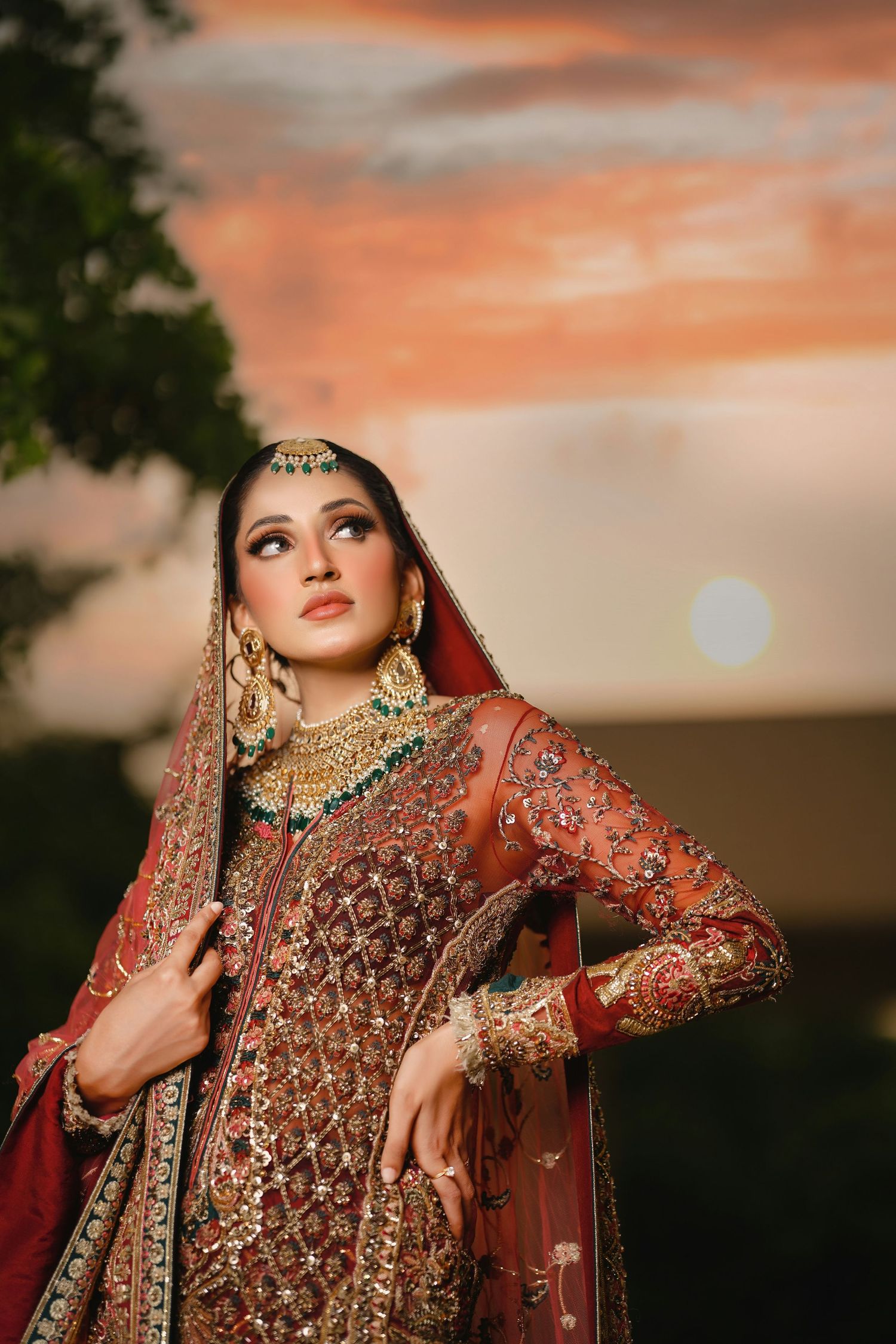 Bride in ornate red and gold embroidered Pakistani bridal lehenga with dupatta at sunset.