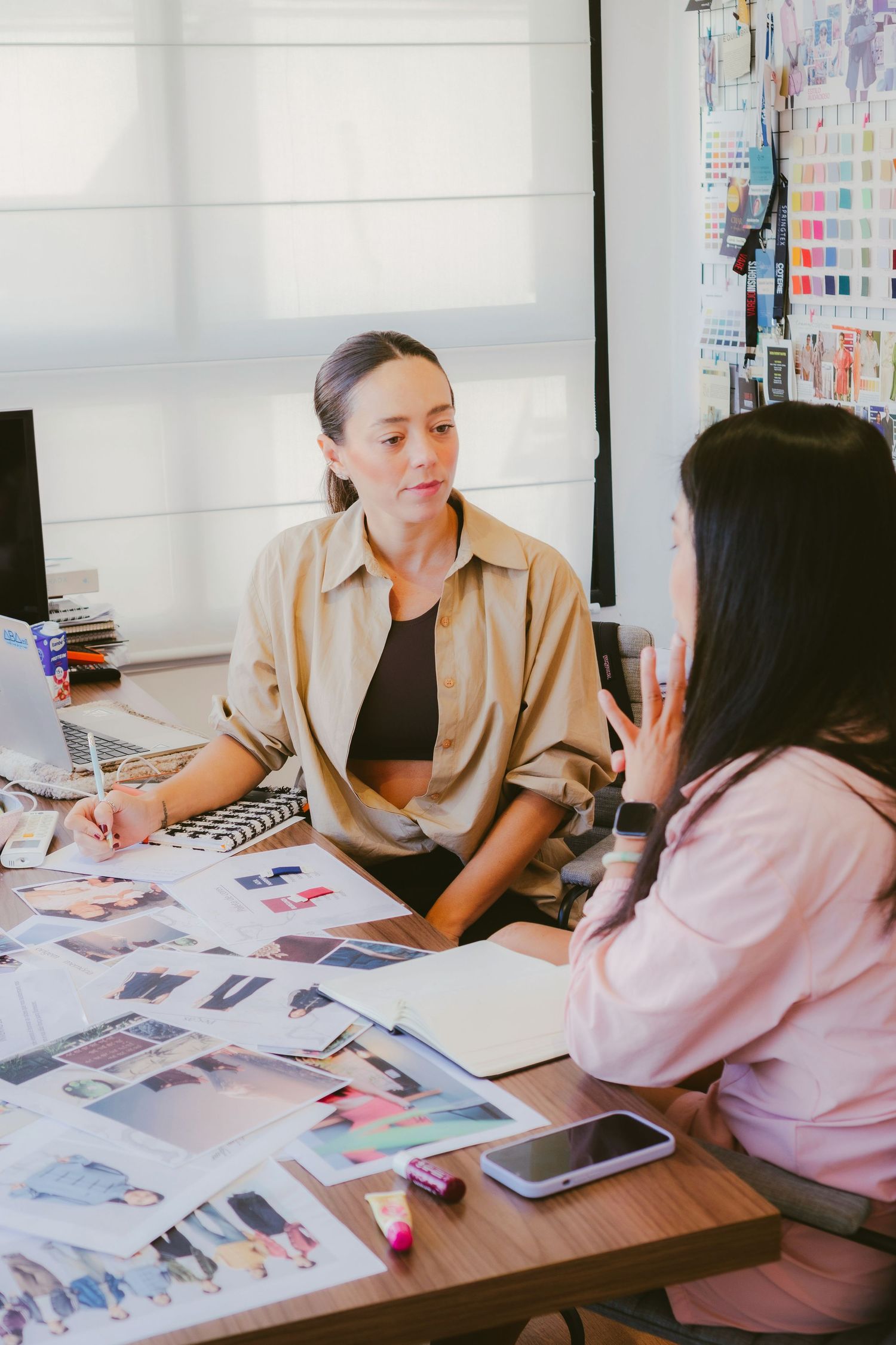Two women discuss fashion designs at a desk covered with mood board images in a bright creative studio.