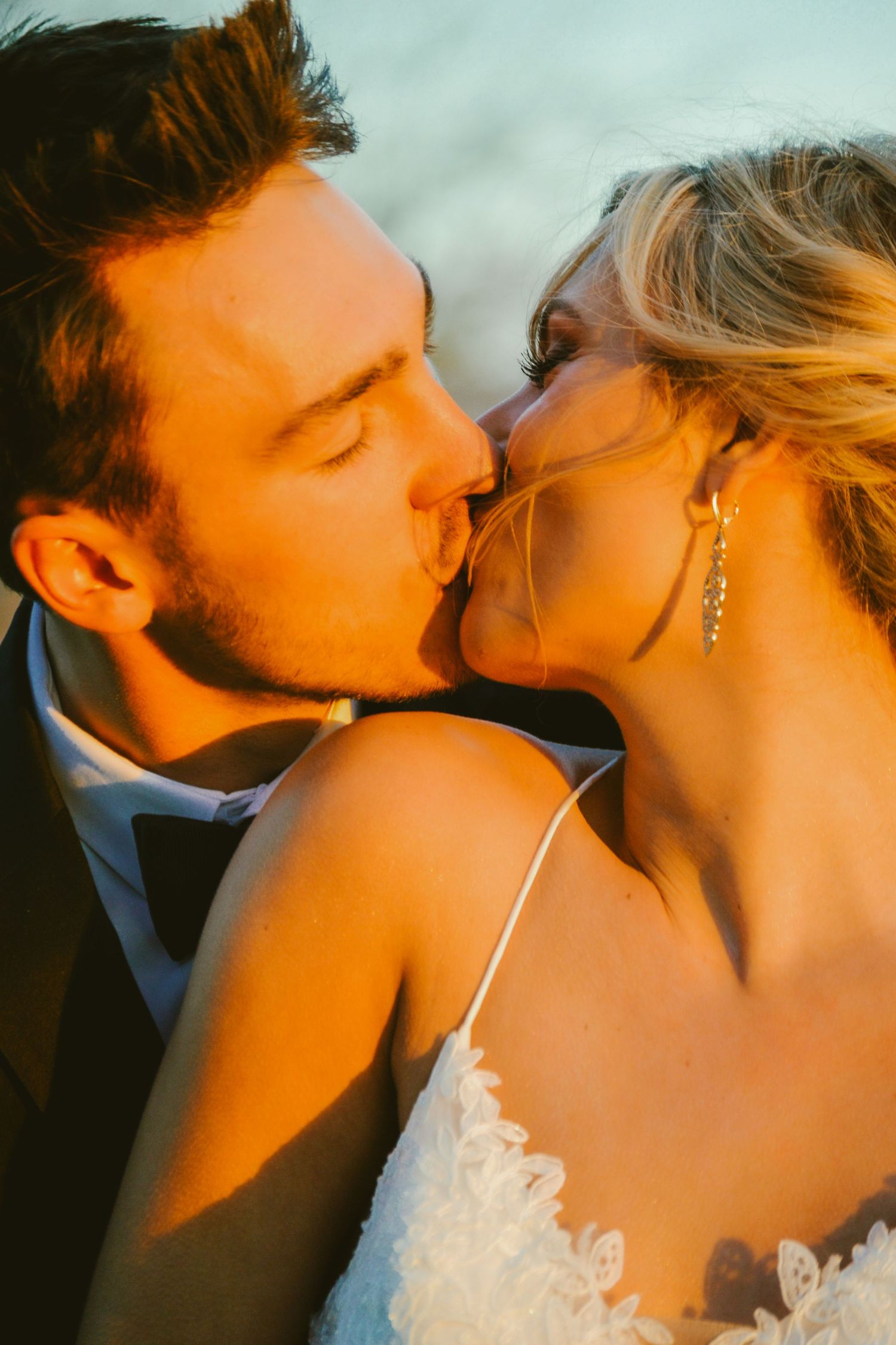 A bride and groom share a romantic kiss in golden sunset light, the bride wearing a lace dress and crystal earrings.