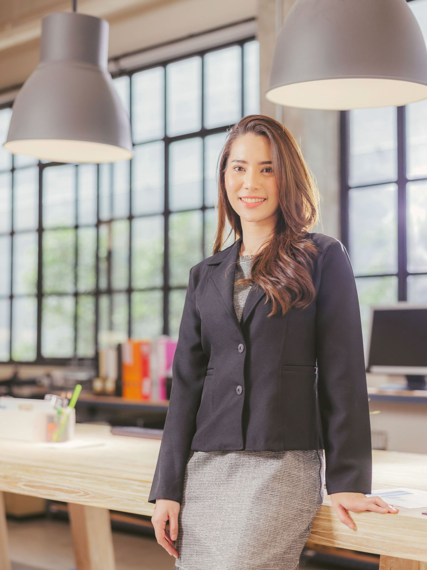 Professional businesswoman in navy blazer and gray skirt smiling confidently in a modern office workspace.