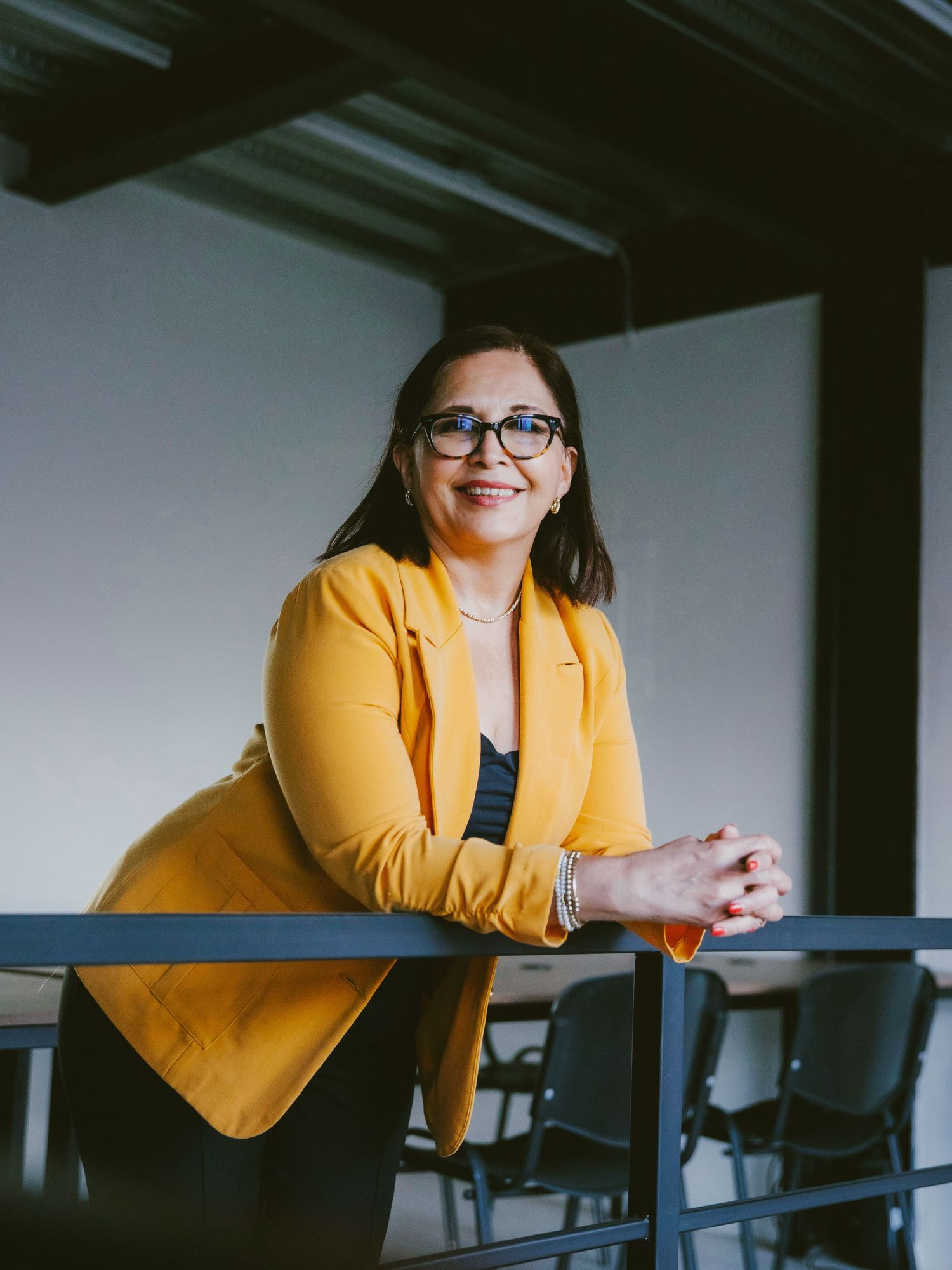 Woman in yellow blazer and glasses smiles while leaning on a railing in a modern office space.