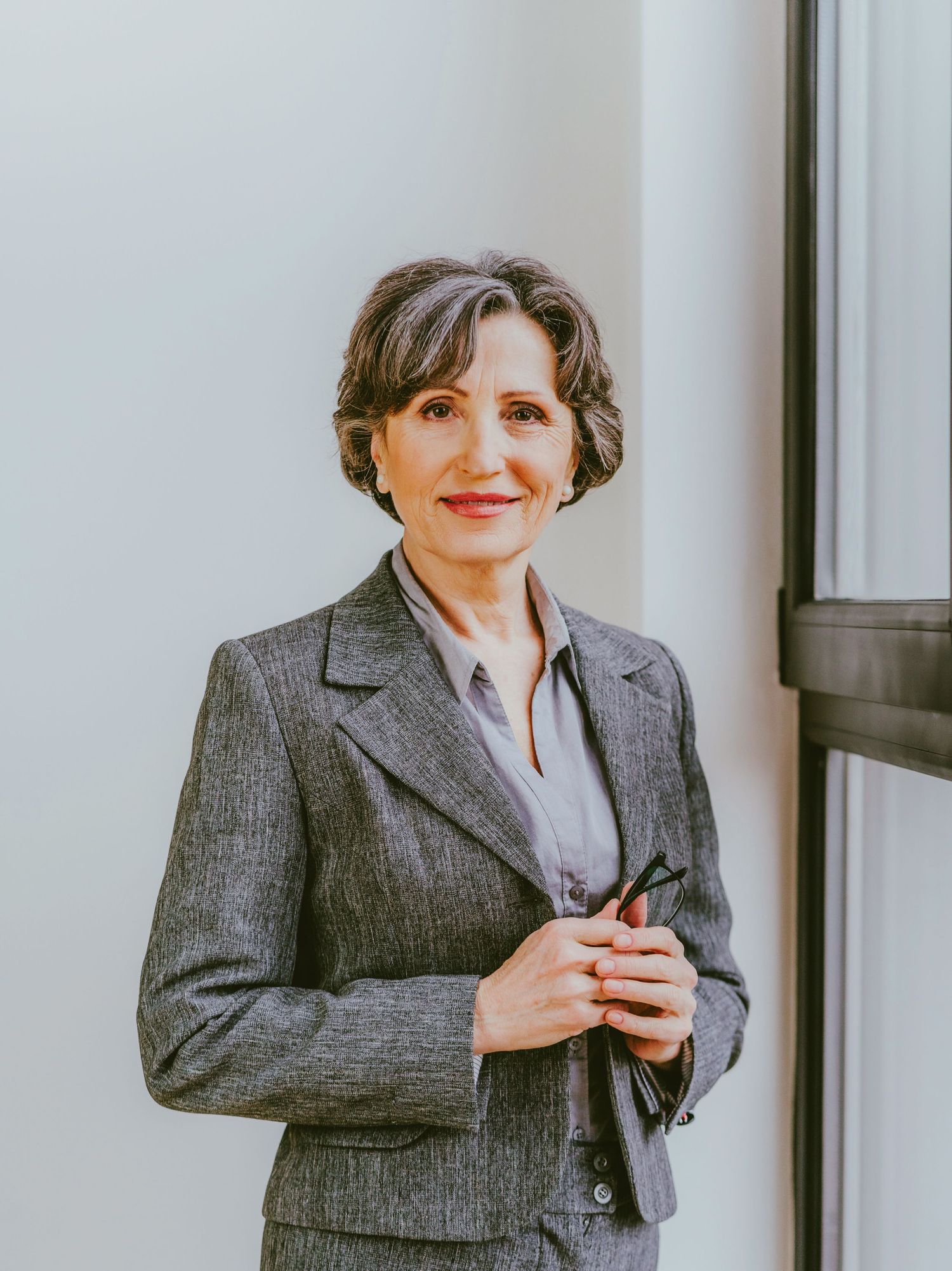 Professional businesswoman in gray suit smiling confidently, holding glasses, standing near window in modern office.