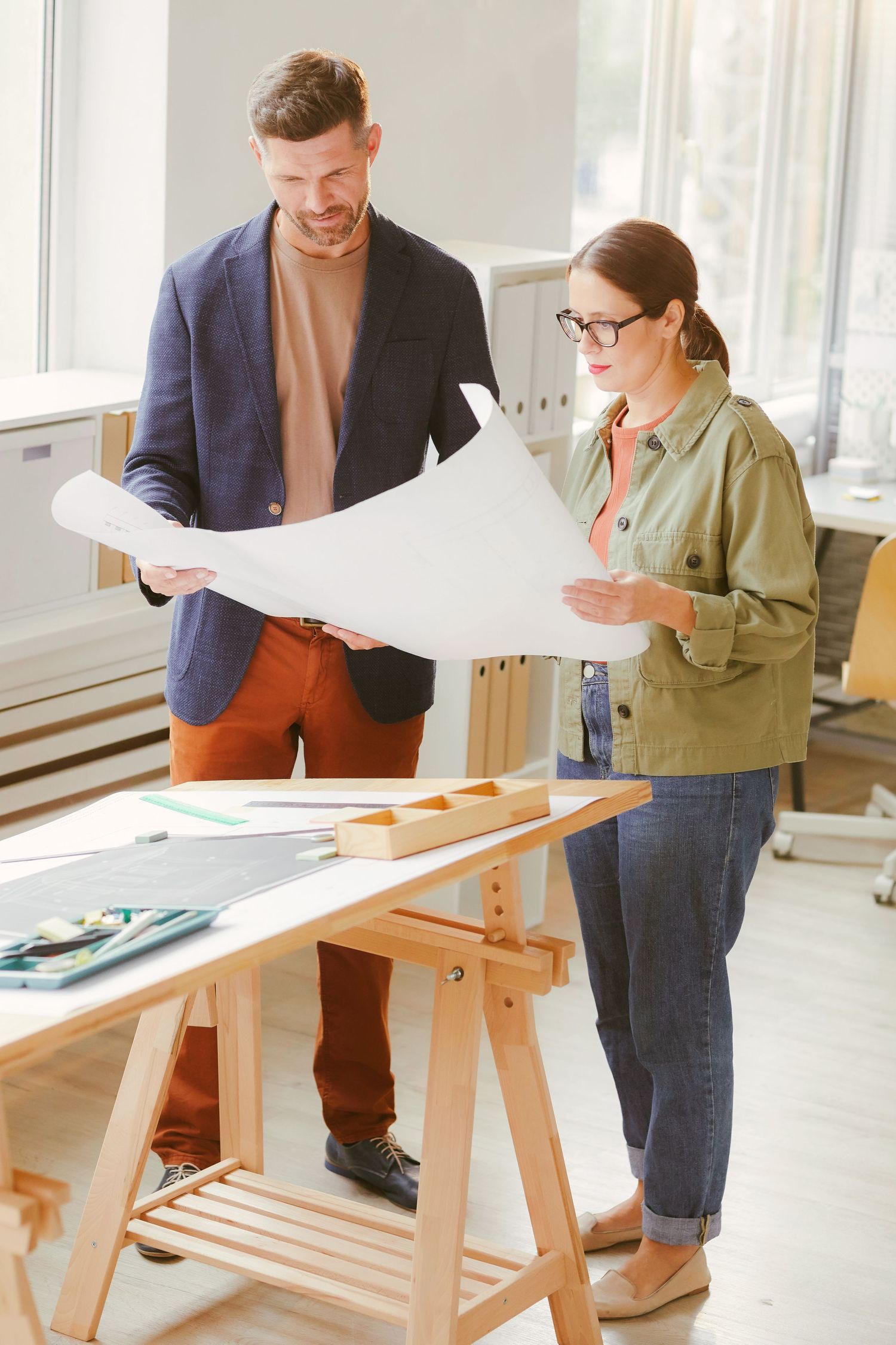 Two professionals reviewing architectural blueprints together at a wooden drafting table in a bright modern office.