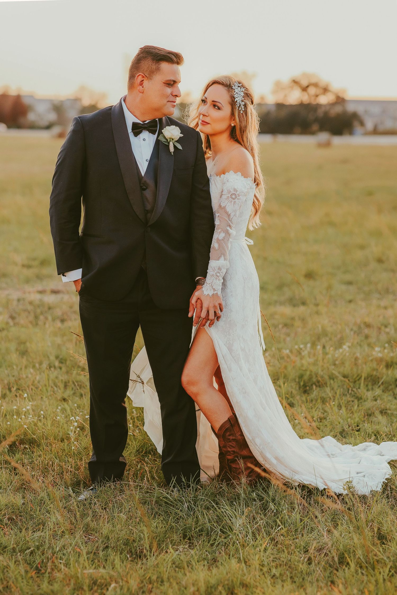 Bride in lace off-shoulder gown with cowboy boots and groom in black tuxedo pose in golden hour field.