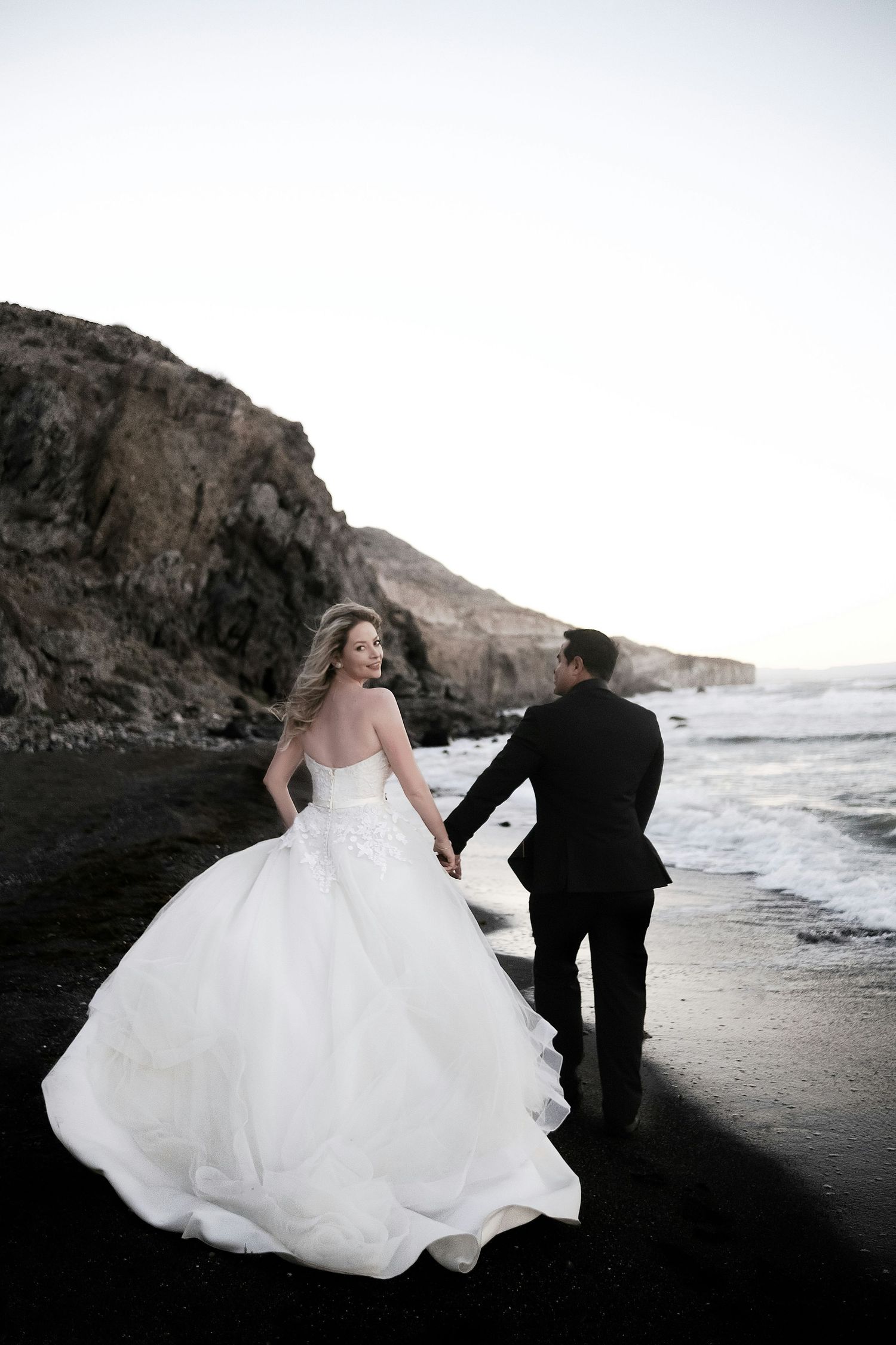 Bride in flowing white ballgown walking with groom on dramatic black sand beach at dusk.