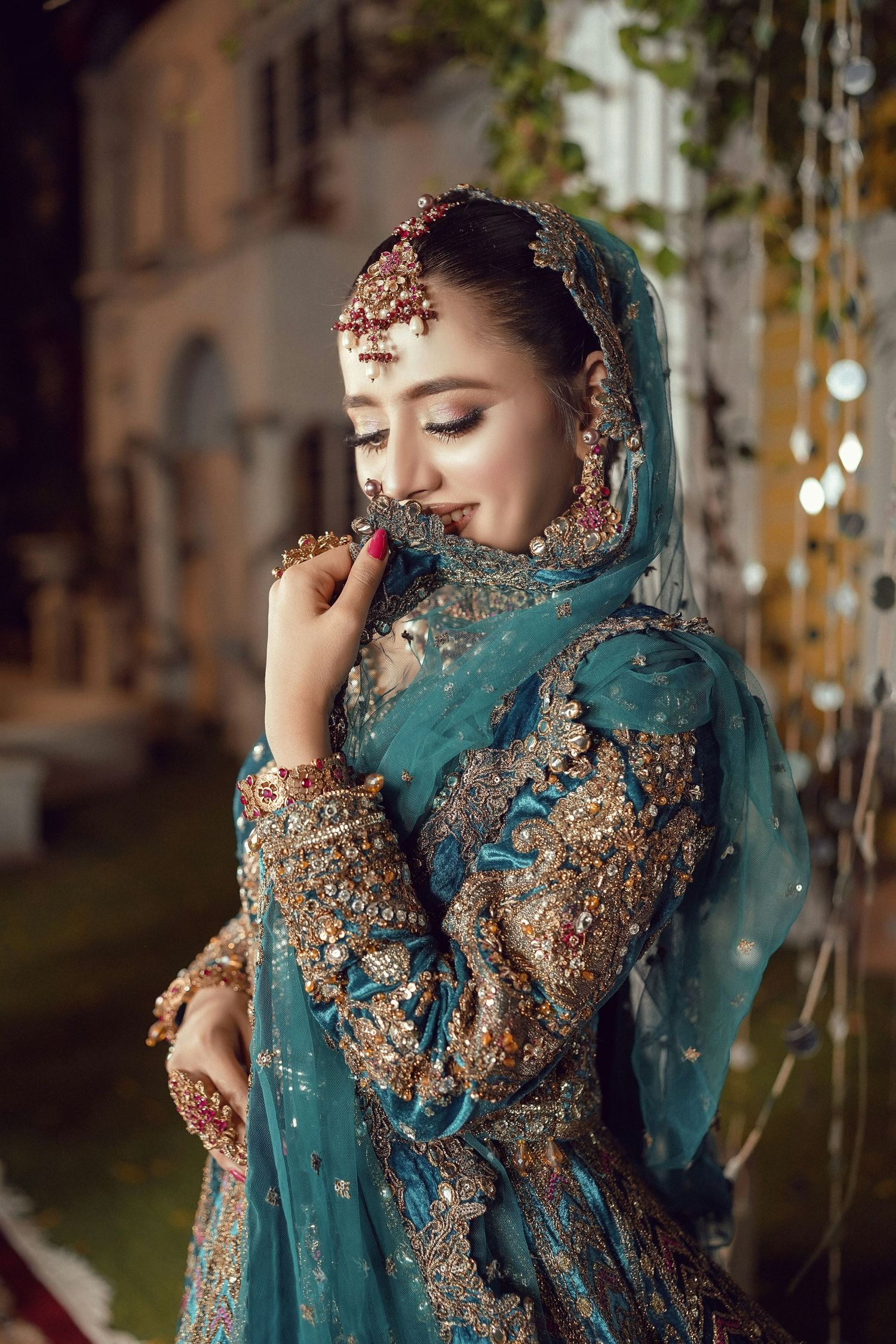 Beautiful bride in teal embroidered Pakistani bridal lehenga with gold jewelry smiling softly.