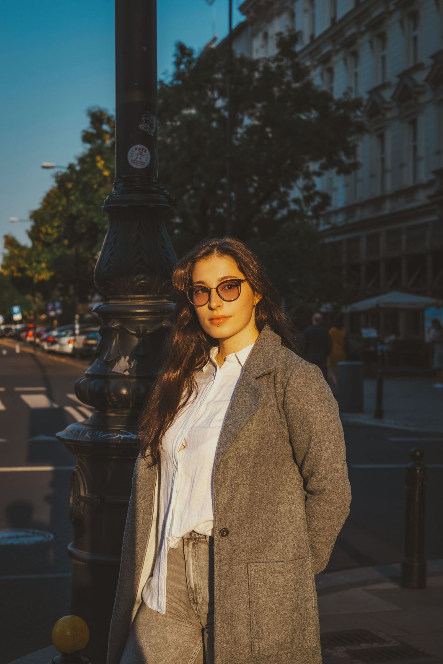 Young woman in gray coat and sunglasses leaning on lamppost on sunny city street.