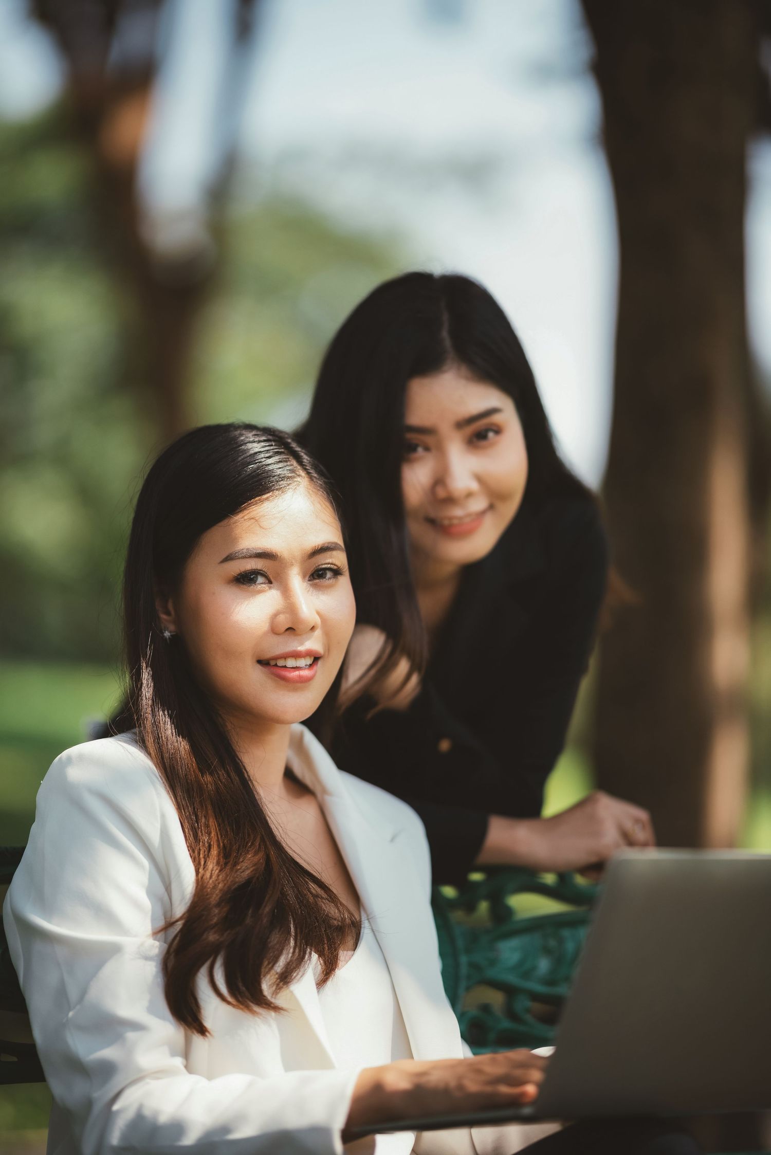 Two Asian businesswomen collaborating on laptop outdoors in a sunny park setting.