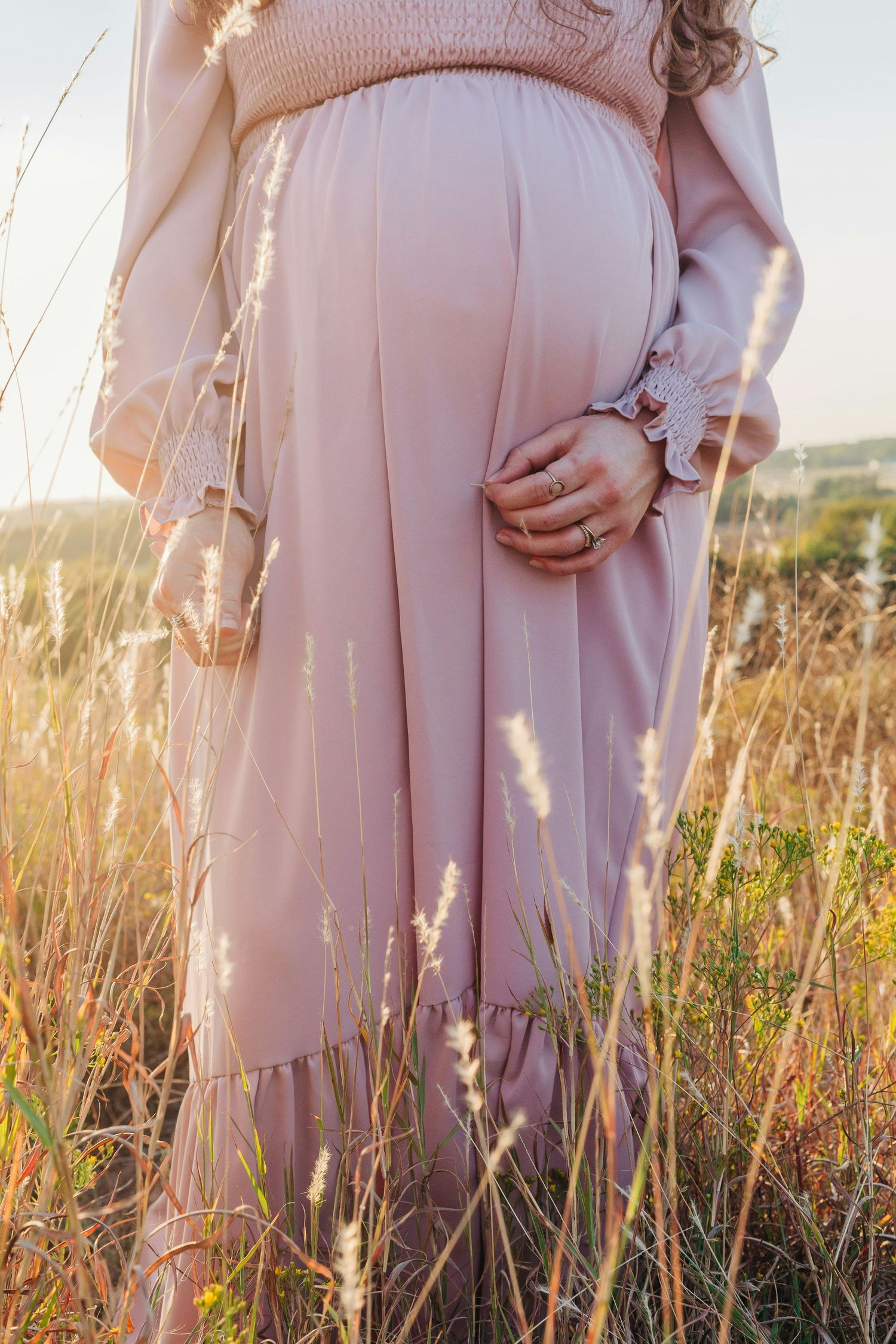 Pregnant woman in pink maxi dress holding baby bump in golden sunlit meadow at sunset.