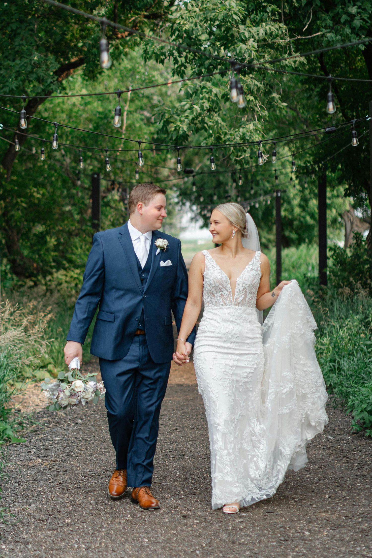 Smiling bride and groom walk together under string lights on a garden path, bride in lace gown, groom in navy suit.