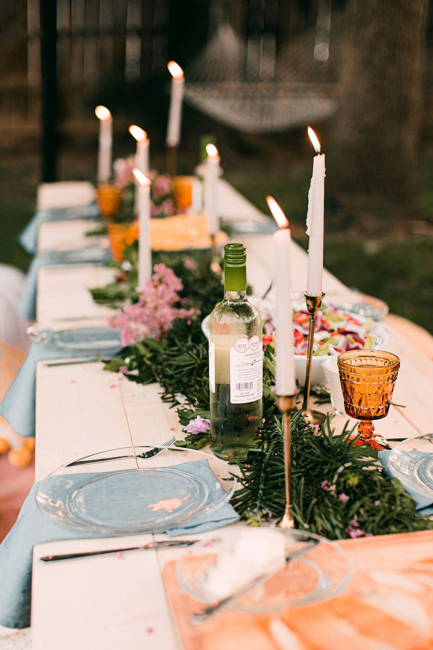 Outdoor garden dinner table set with candles, wine bottle, blue plates, and greenery runner at dusk.