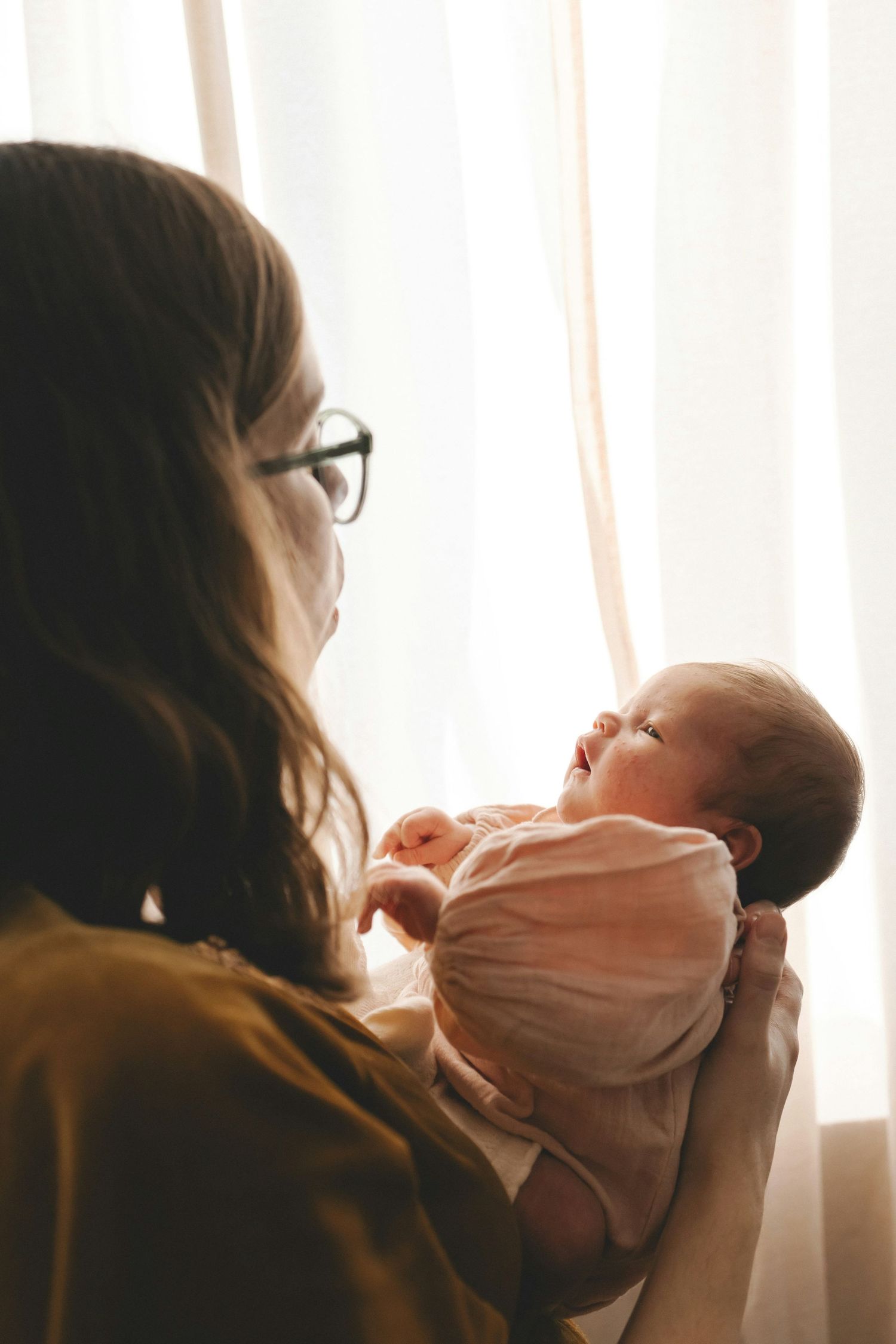 Mother with glasses holding newborn baby up near sheer white curtains in warm natural light.