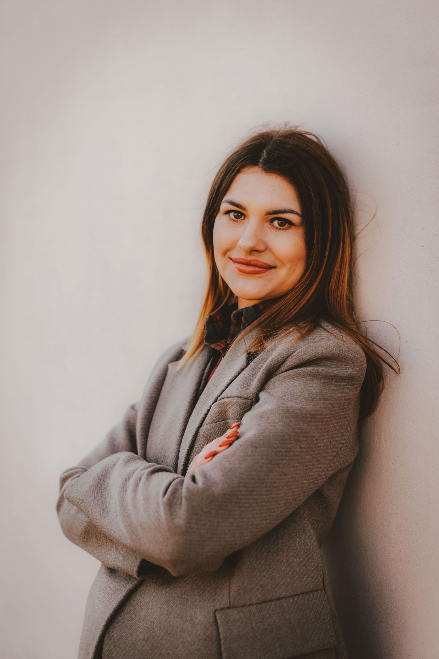 Confident young brunette woman in gray blazer smiling with arms crossed against white wall.