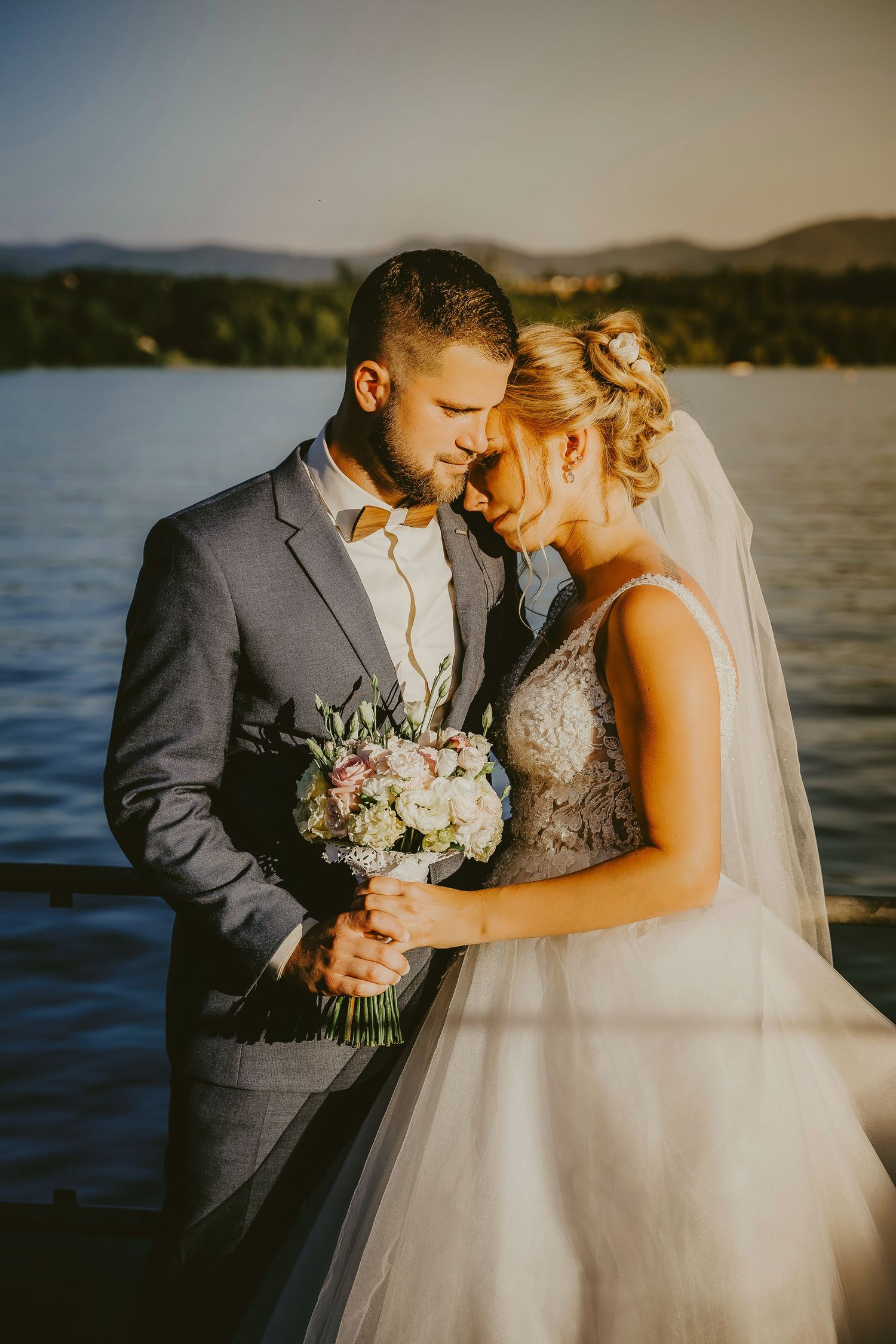 Bride and groom share a romantic moment by a lake, she holds a pastel bouquet, both in elegant wedding attire at sunset.