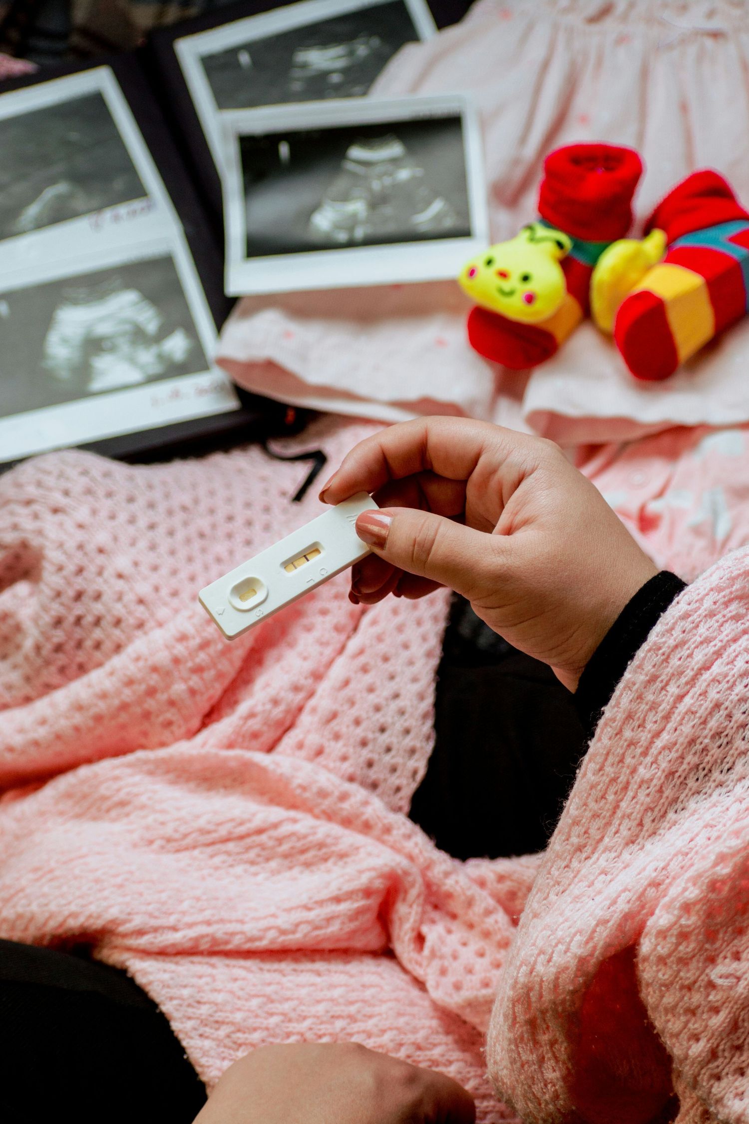 Woman holding positive pregnancy test surrounded by ultrasound photos, baby clothes, and toys on pink blanket.