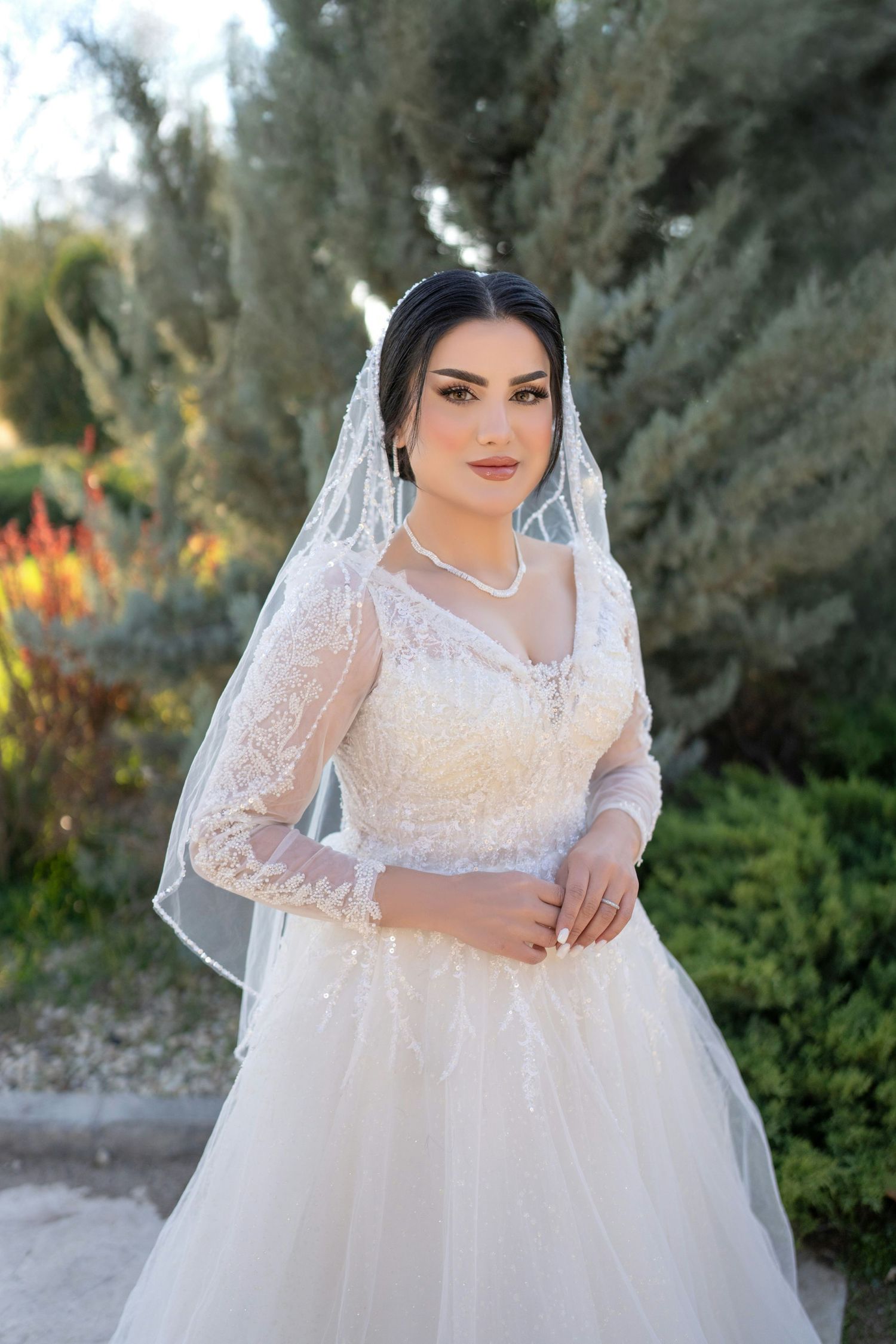 Bride in long-sleeve white lace wedding gown with veil posing in a garden.