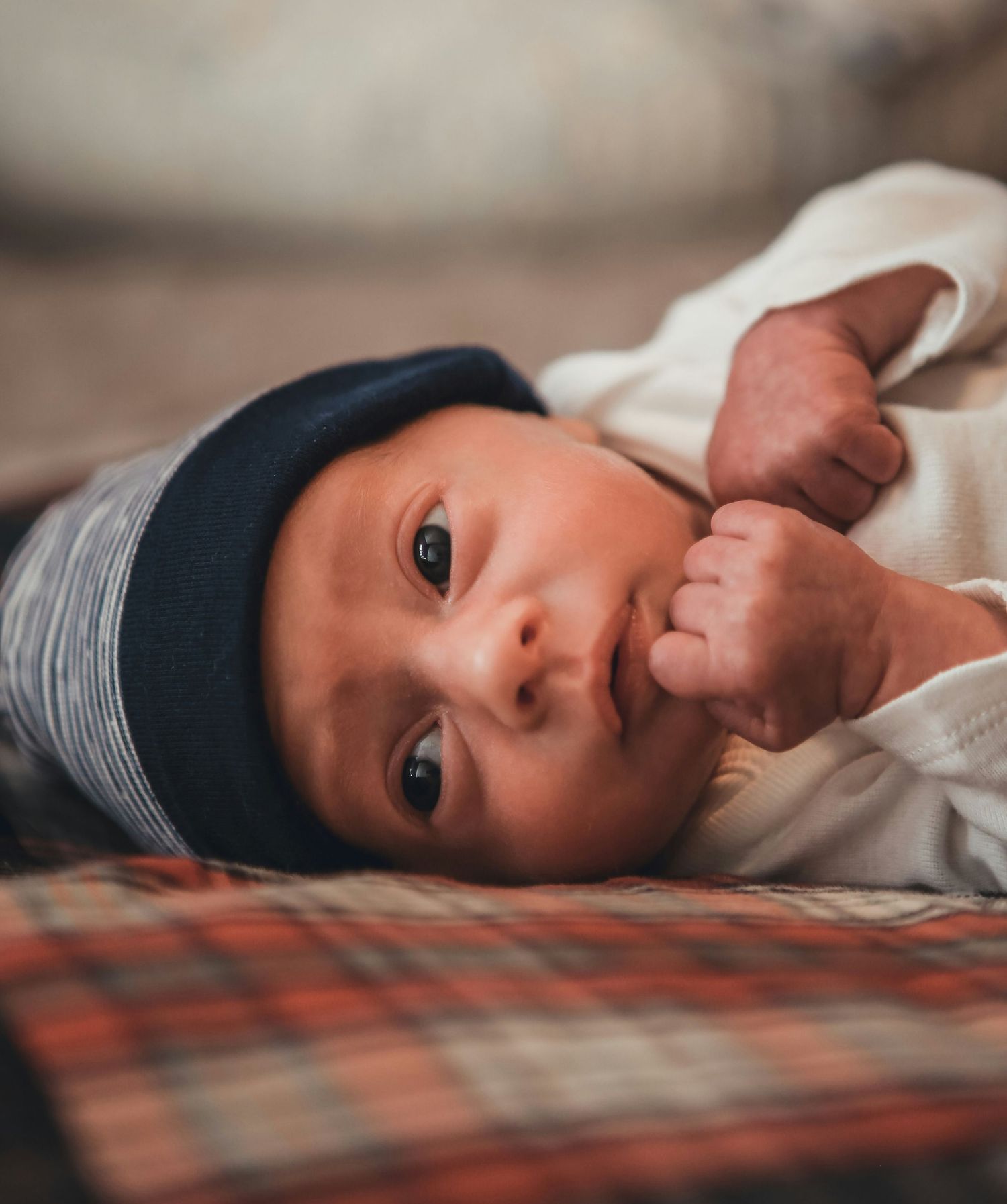 Wide-eyed newborn baby in dark beanie hat lying on plaid blanket, close-up portrait.