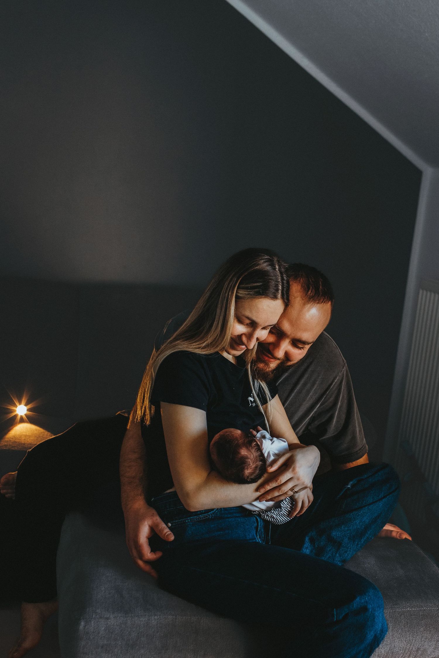 A couple sits together in a dimly lit room, lovingly gazing at their newborn baby held in their arms.