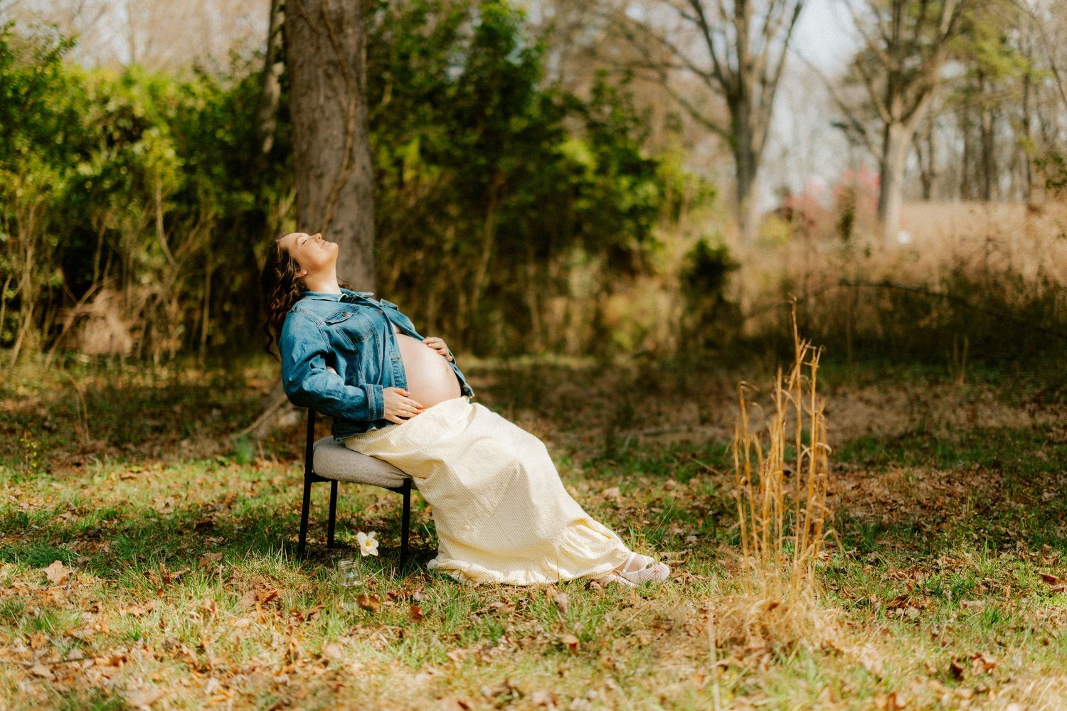 Pregnant woman in denim jacket and white skirt sitting on chair in a sunlit outdoor garden setting.