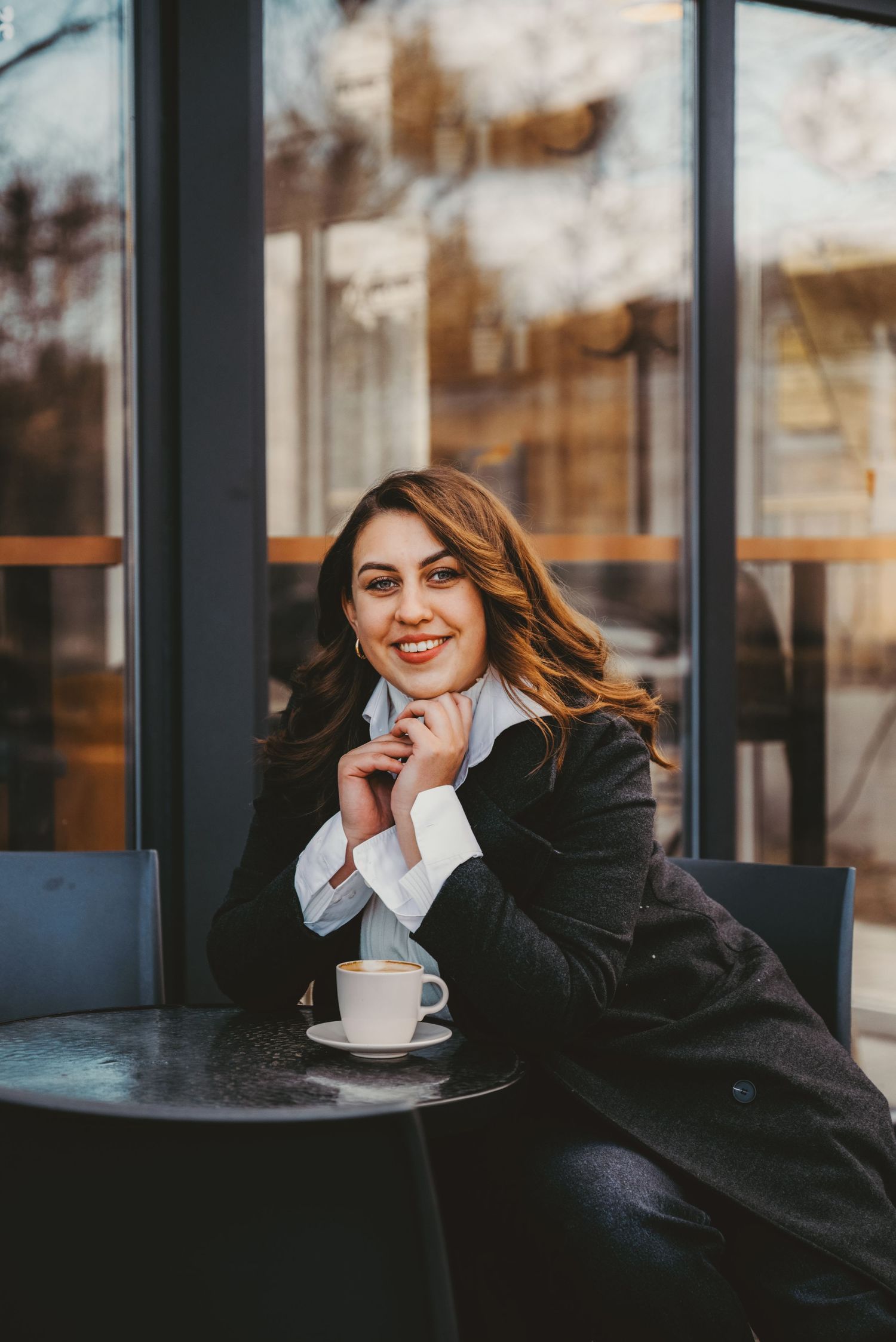 Elegant woman in black coat sitting at cafe table with coffee cup smiling.