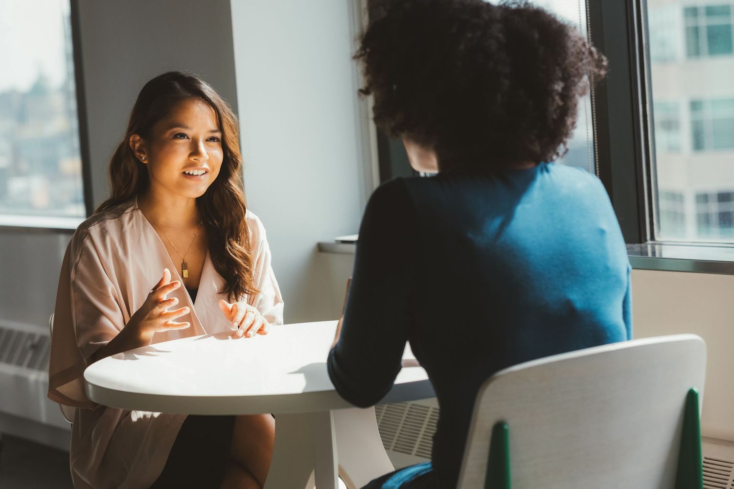 Two professional women having a business meeting at a white table in a modern office with large windows.