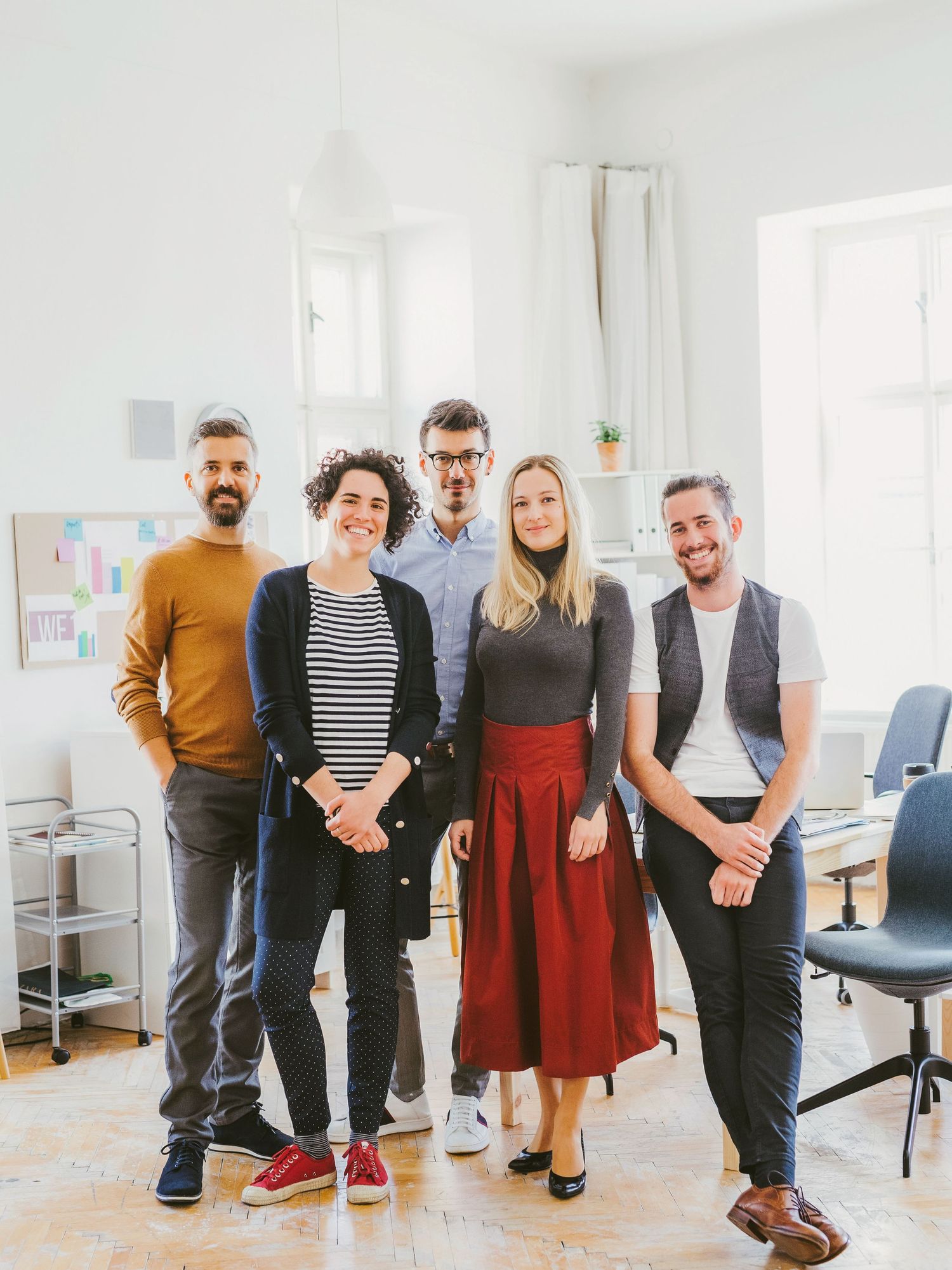 Five smiling colleagues pose together in a bright modern office workspace for a team photo.