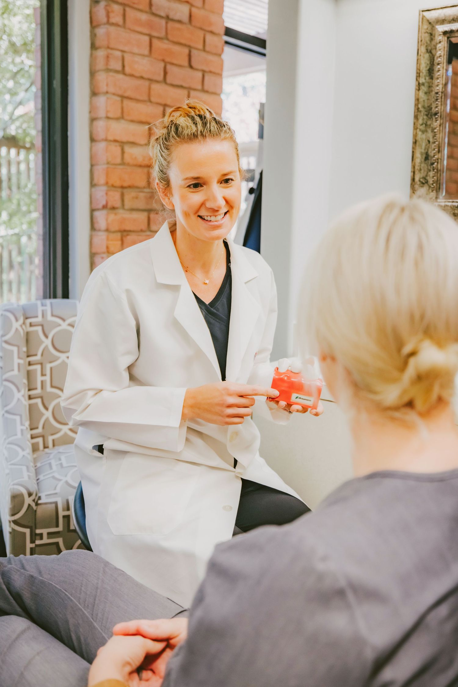 A smiling female dentist in a white coat holds a dental model while consulting with a patient in a modern office.