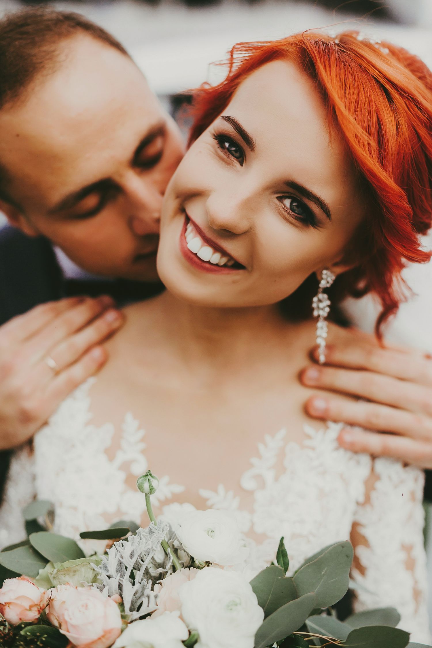 Smiling bride with red hair in white lace dress holds bouquet as groom kisses her cheek on wedding day.