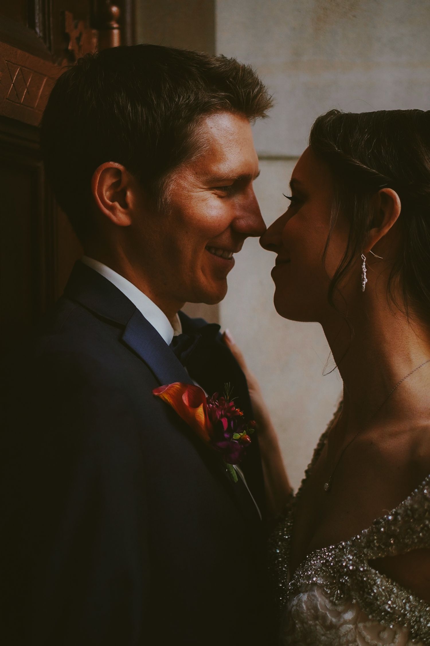 A bride and groom share an intimate moment, nearly kissing, dressed in wedding attire with a floral boutonniere.