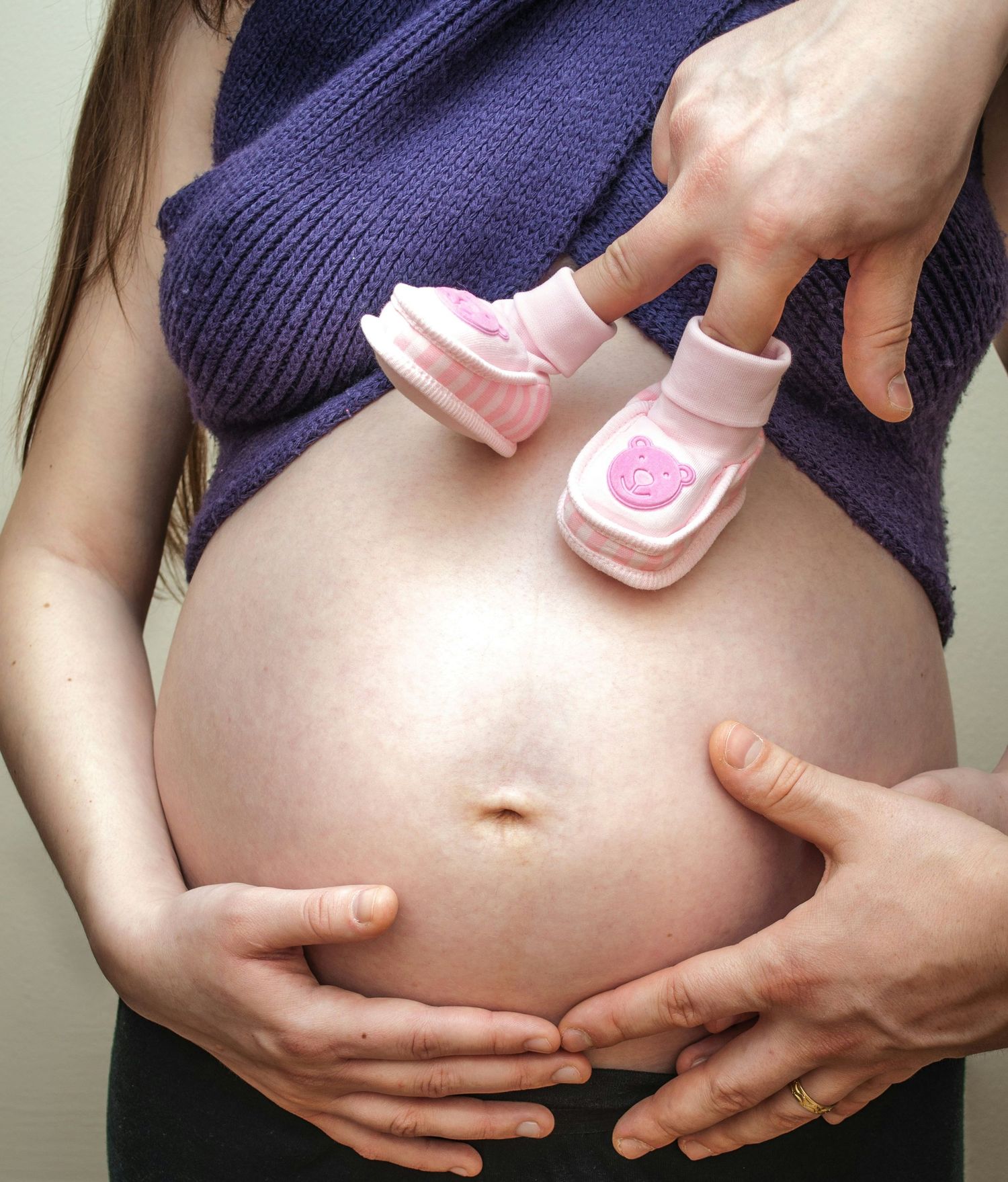 Pregnant woman holding pink baby booties on her bare belly, with partner's hands cradling bump.