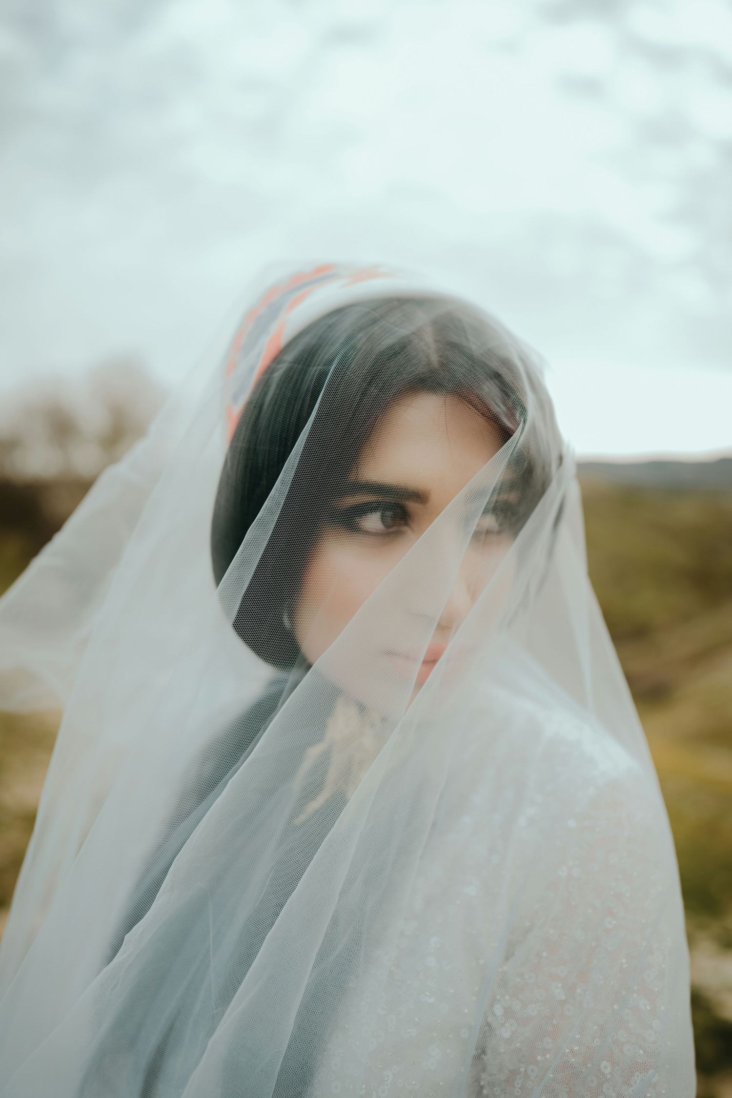 Artistic portrait of dark-haired bride with sheer veil draped over face against cloudy sky.