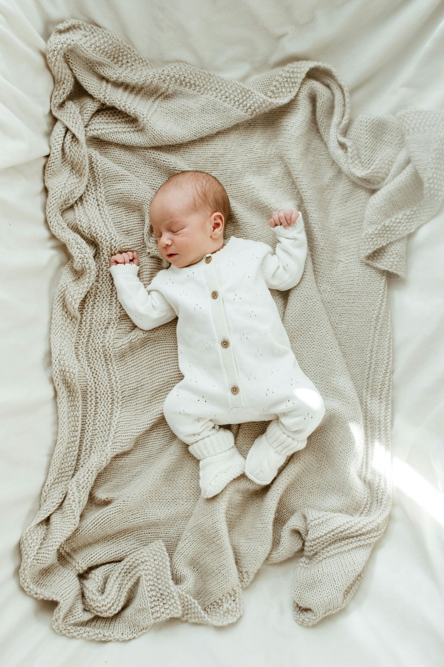 Peaceful newborn baby sleeping in white knit outfit on a beige textured blanket.