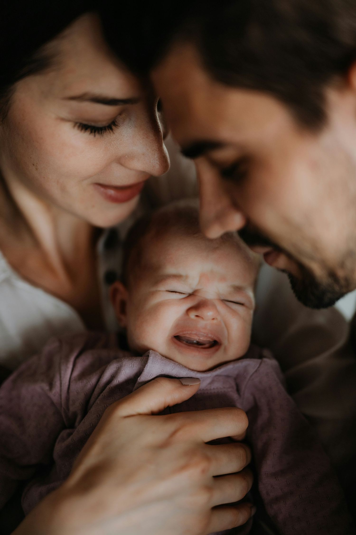 Parents smiling tenderly at crying newborn baby dressed in purple onesie, close-up family portrait.