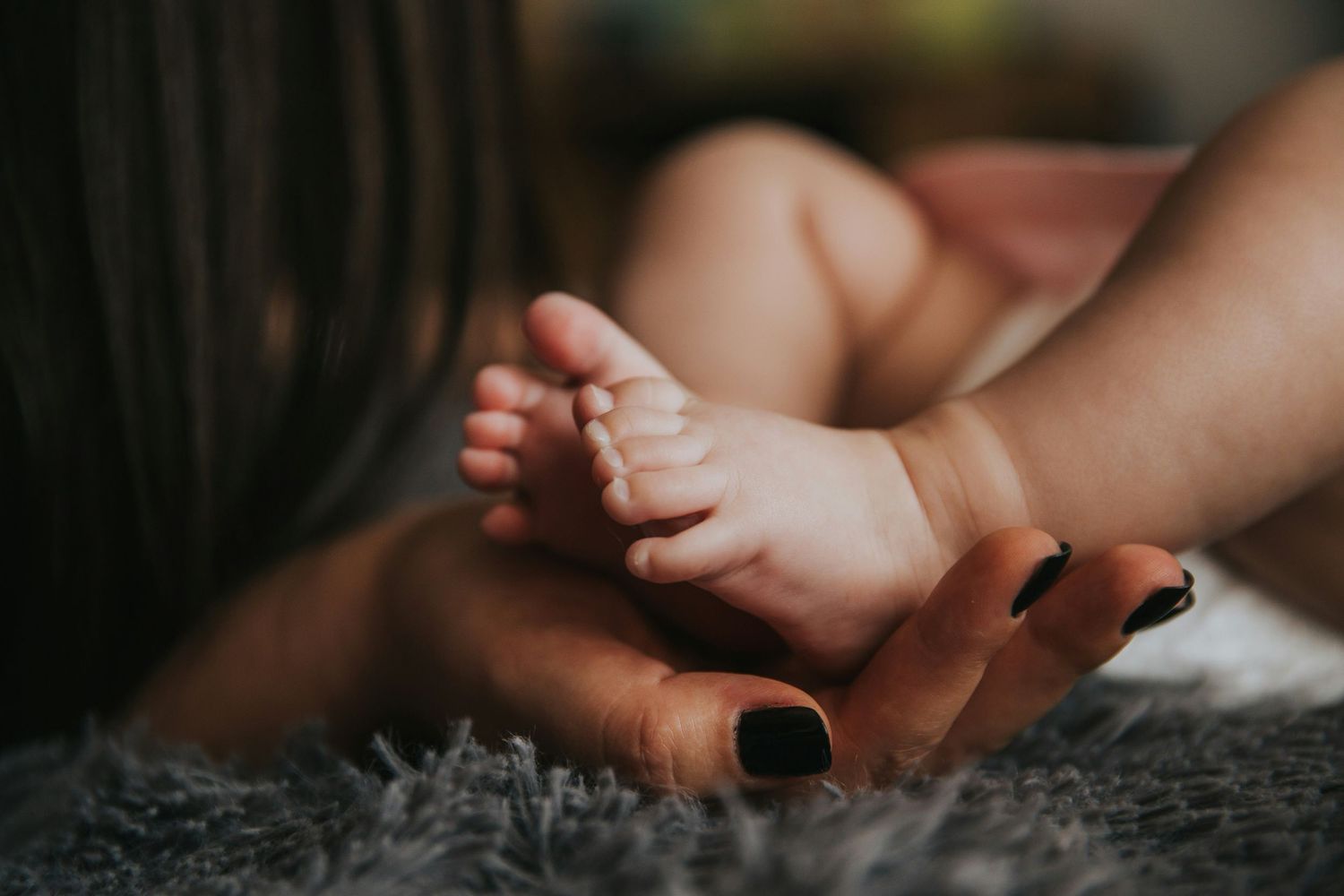 Close-up of newborn baby feet cradled gently in mother's hands with dark painted nails on soft rug.
