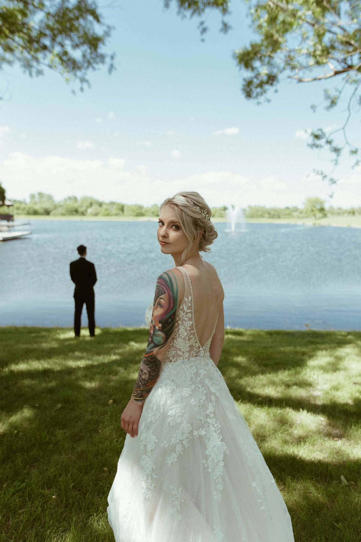 Tattooed bride in backless lace gown stands by lakeside with groom behind her.