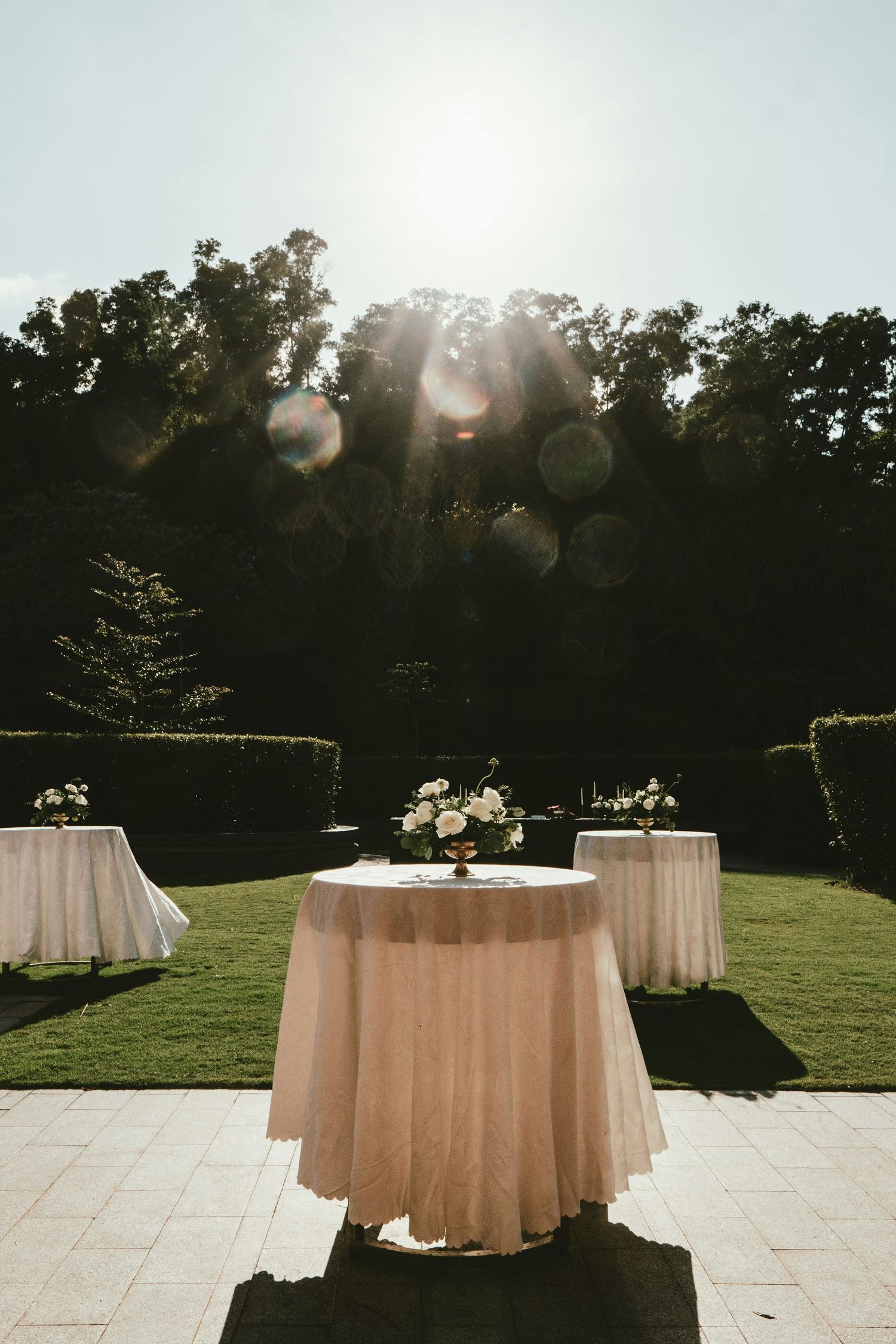 Outdoor wedding cocktail hour setup with white linen tables on stone patio with sun flare and hedges.