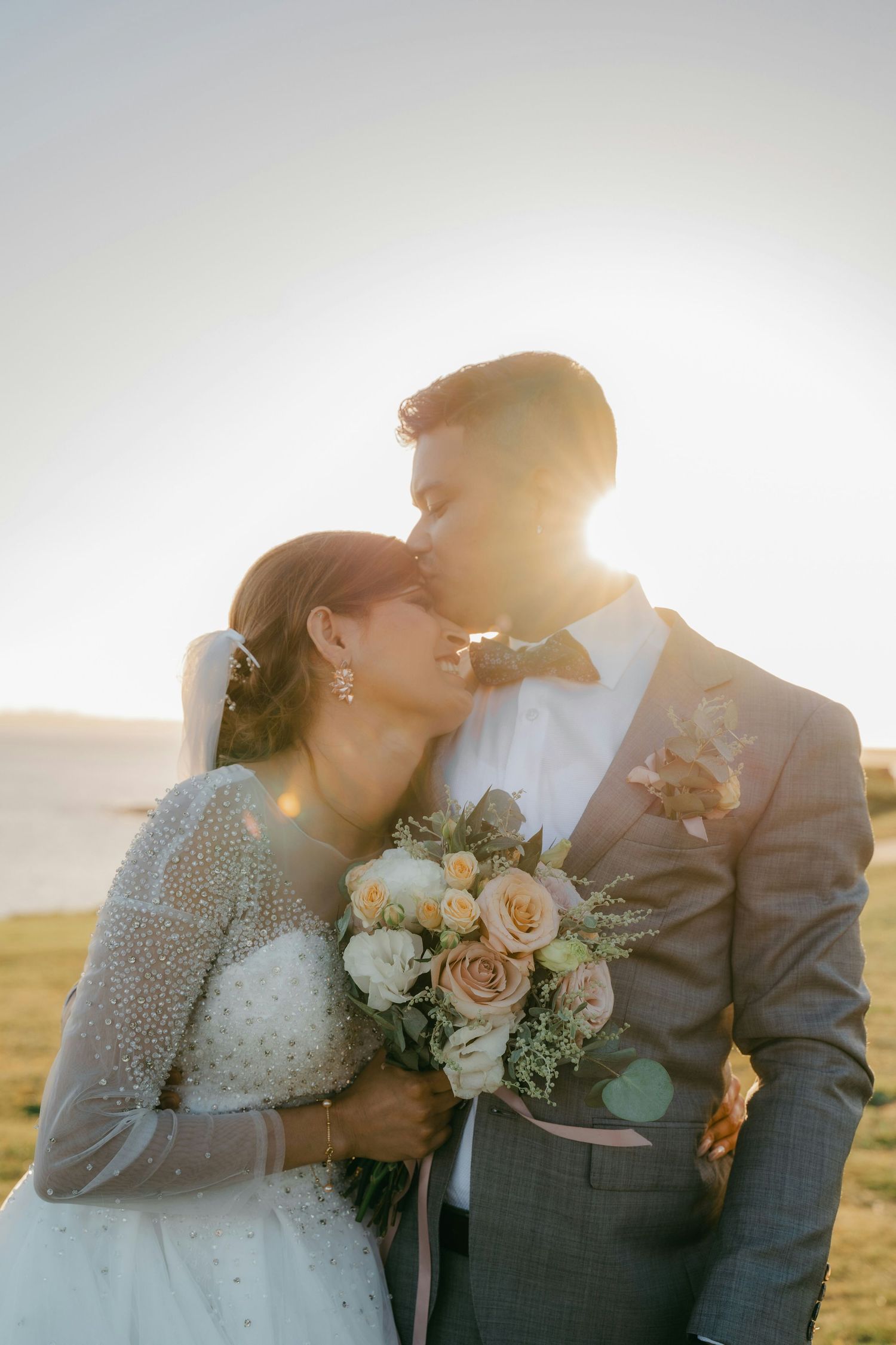 Bride and groom share a tender kiss at sunset by the sea, bride holding a bouquet of peach and white roses.