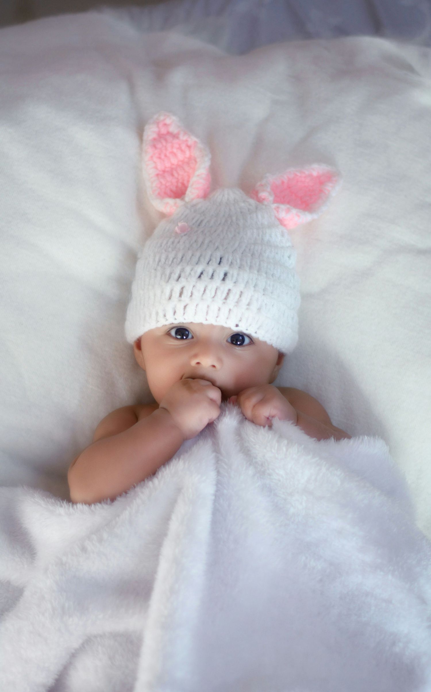 Adorable newborn baby wearing white knit bunny ear hat lying on soft white blanket.
